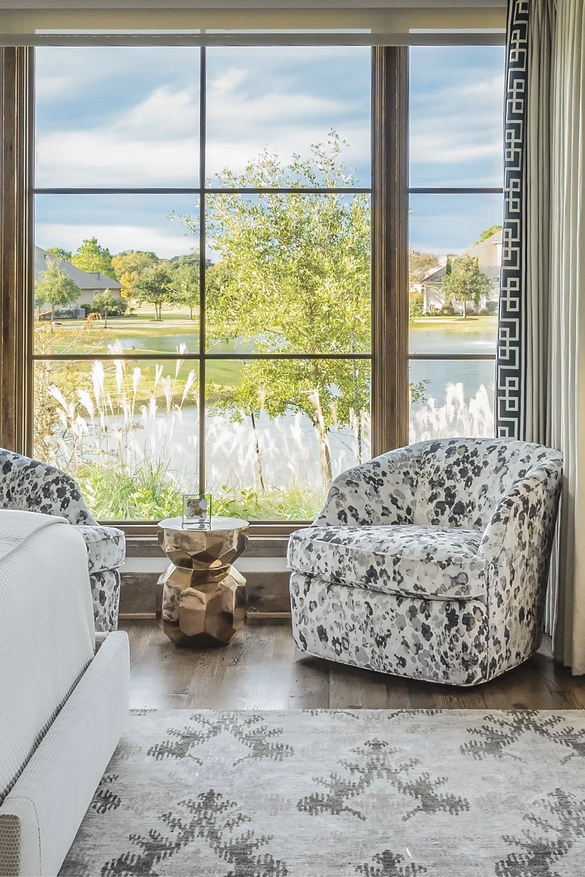 Modern ranch bedroom window seating with patterned chairs, a faceted metal side table and views across the Houston Oaks lake.