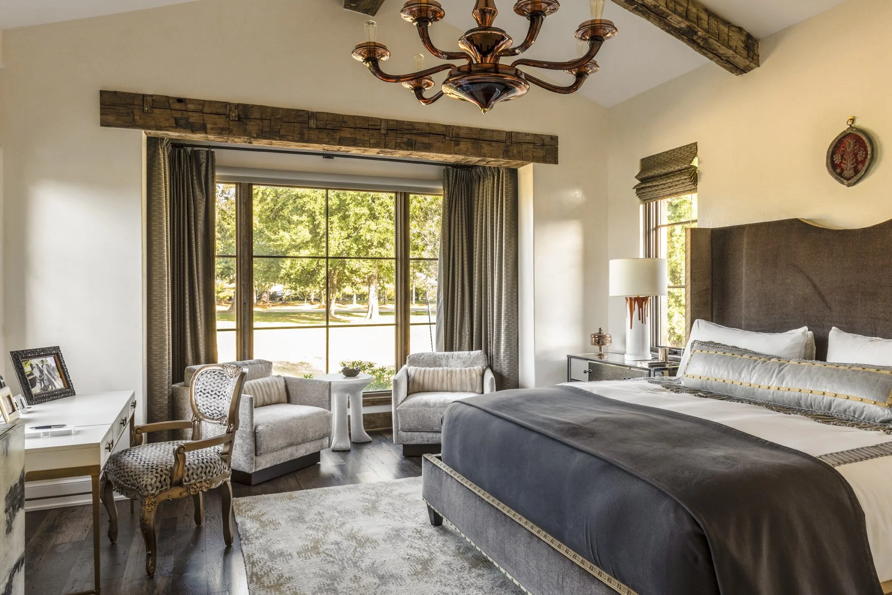 Luxury ranch bedroom at Houston Oaks with a gray upholstered bed, sitting area, reclaimed beams and a large garden window.