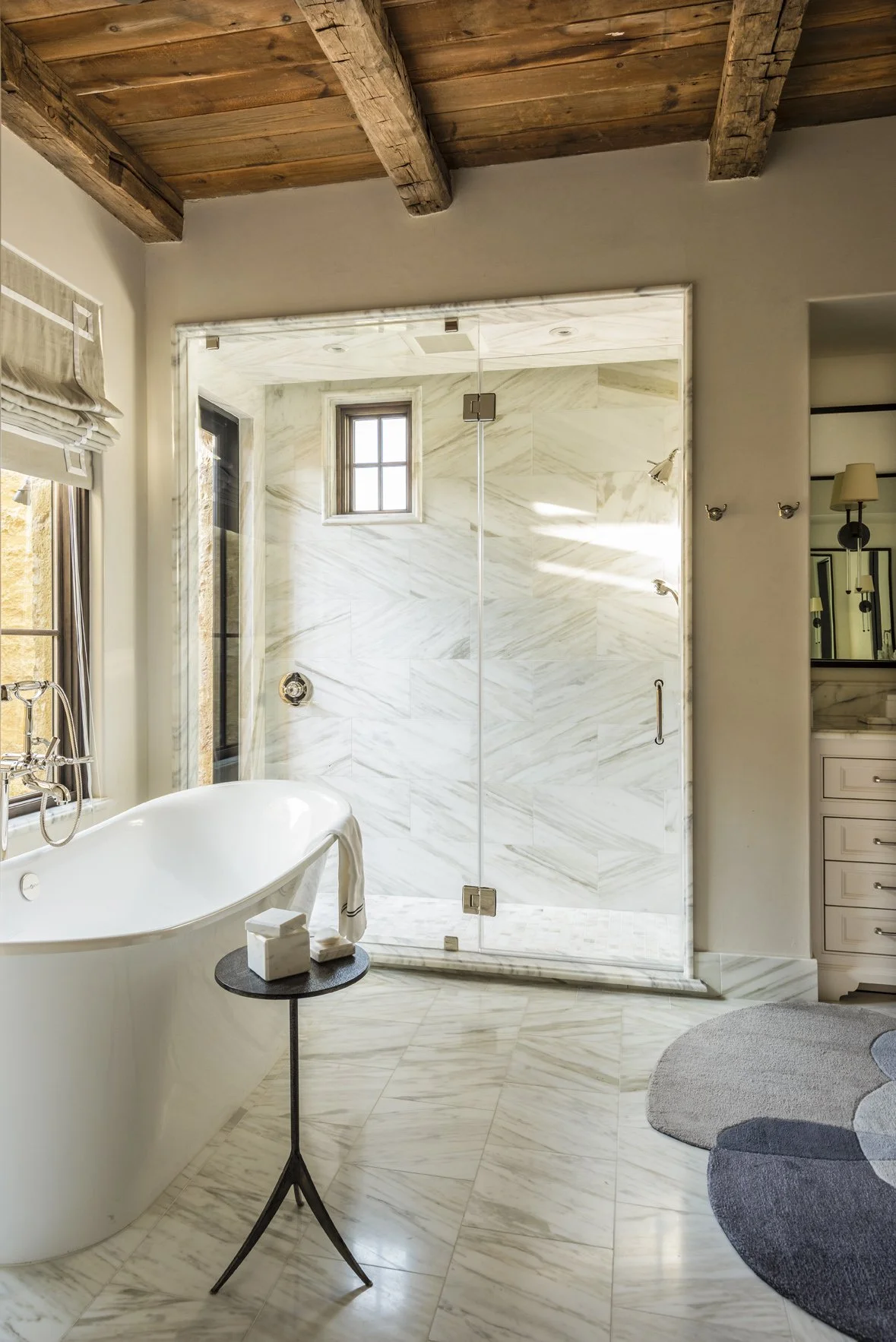 Texas ranch bathroom with a freestanding tub, marble-lined glass shower, reclaimed timber ceiling and sculptural gray rug.