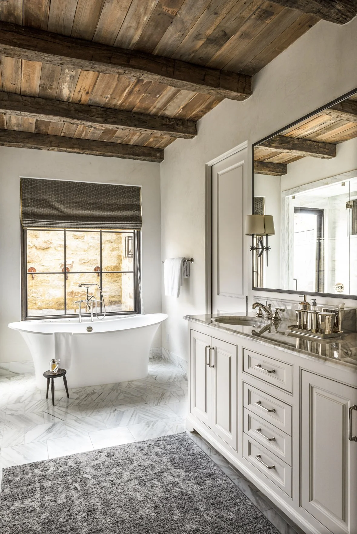 Modern ranch bathroom with a freestanding tub, pale marble vanity, gray rug and a warm reclaimed wood plank ceiling.