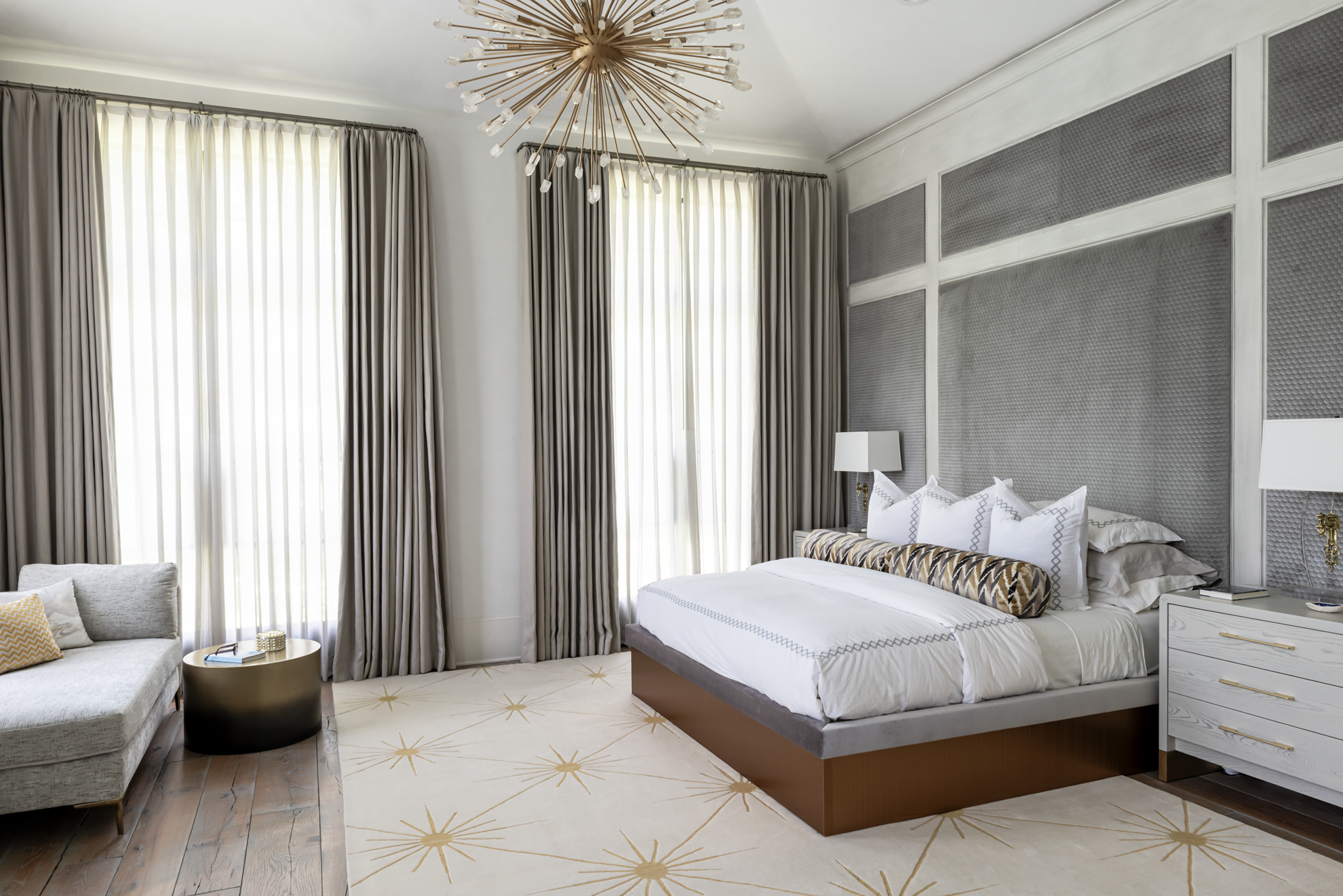 French country home in Houston primary bedroom with a grey upholstered wall, brass chandelier and starburst rug.