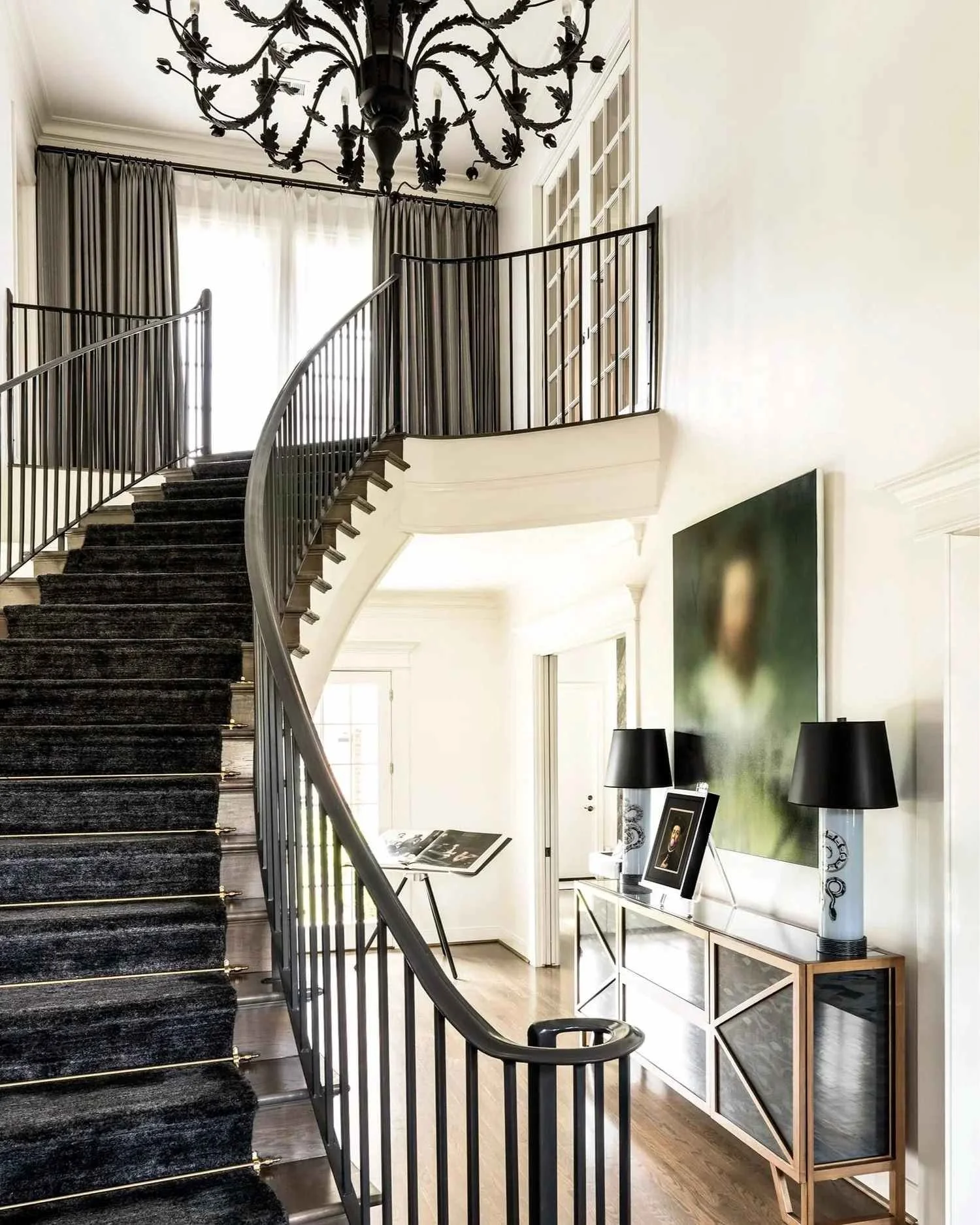 Pastureview residence hallway with a curved staircase, black chandelier, mirrored console and contemporary portrait artwork.
