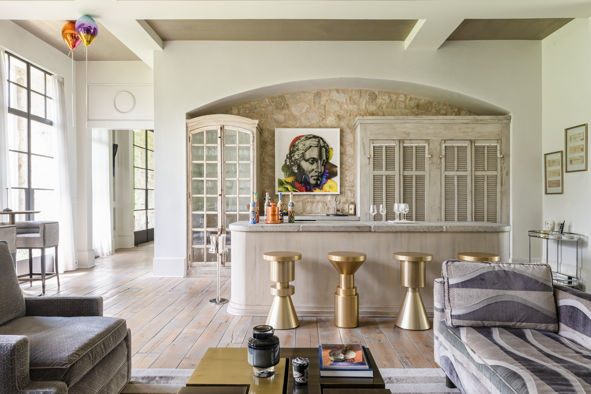 French country home in Houston bar with a stone wall, pale wood cabinetry, brass stools and colourful portrait art.