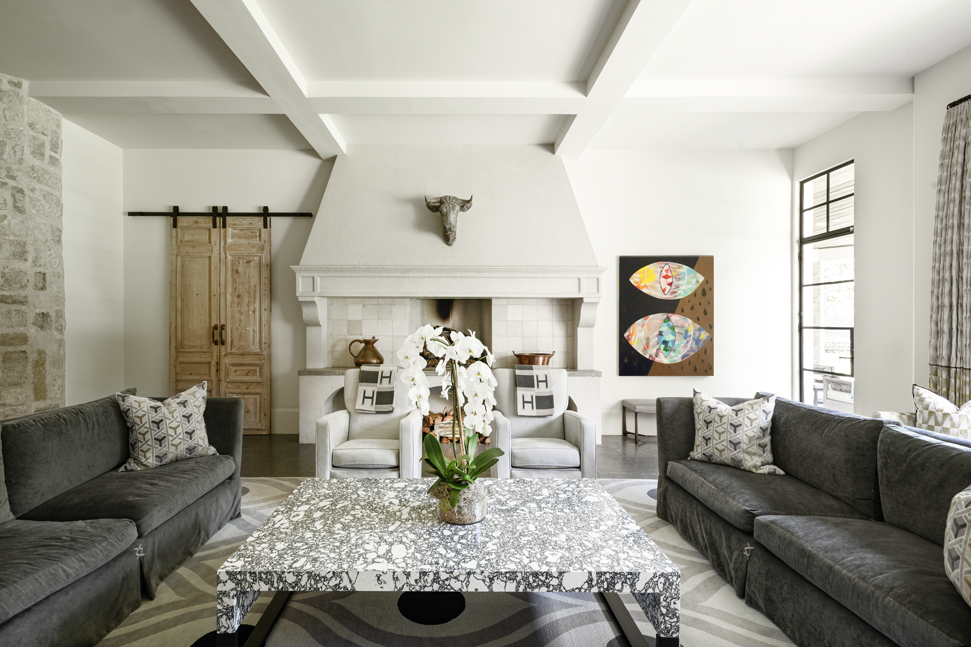 French country home in Houston family room with charcoal sofas, a limestone fireplace, marble table and modern art.