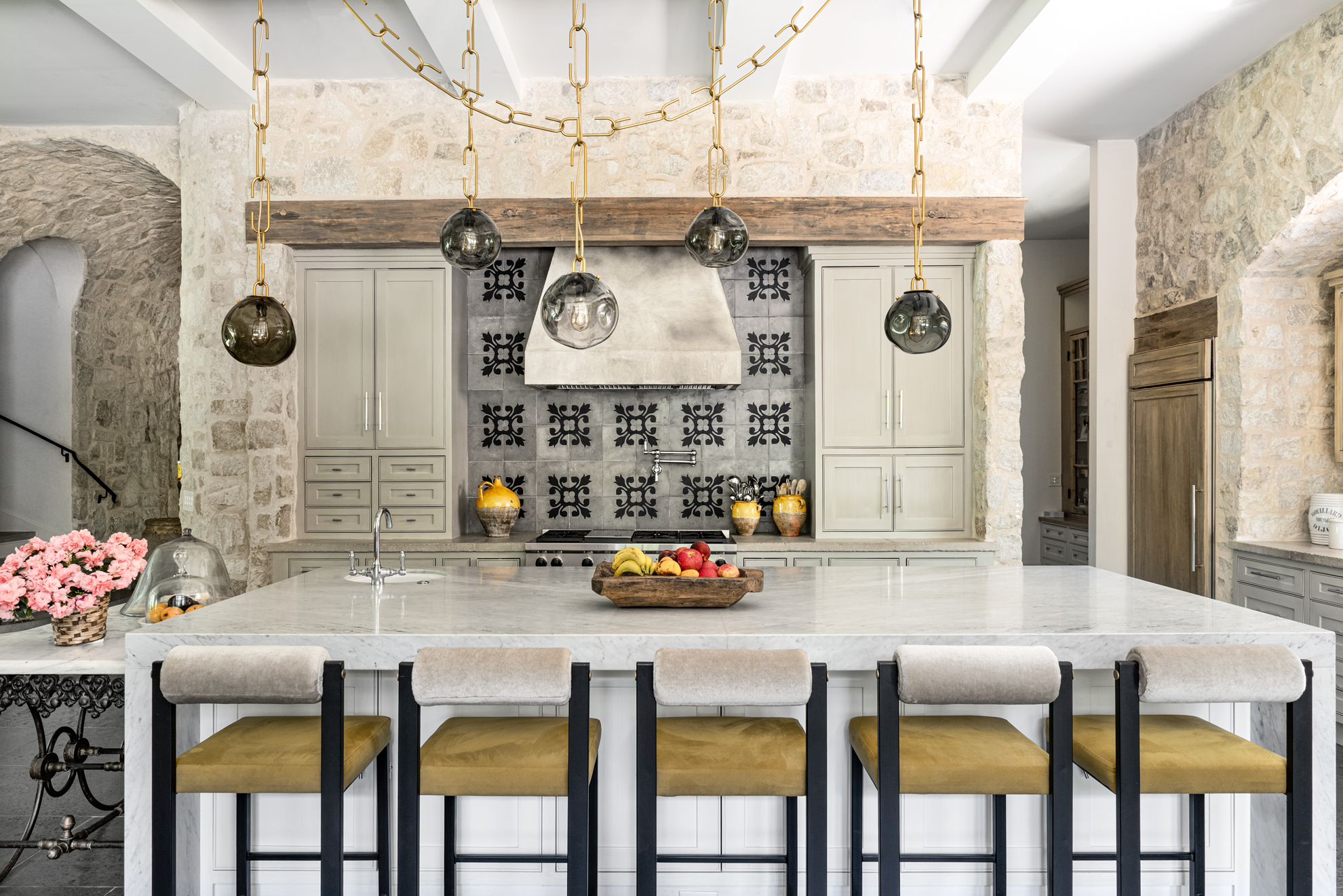 French country home in Houston kitchen with a marble island, mustard stools, patterned backsplash and stone walls.