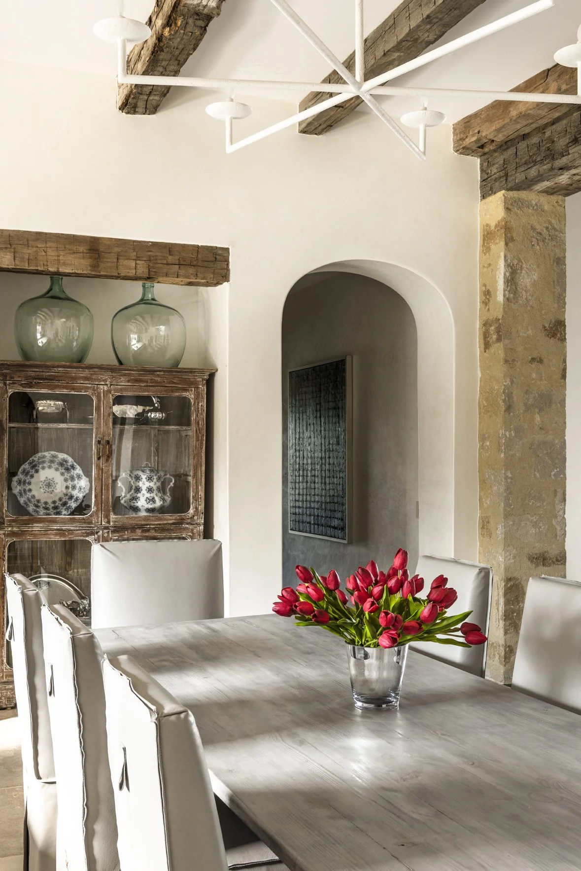 Modern ranch dining room with a gray table, white slipcovered chairs, reclaimed beams, an arched opening and red tulips.
