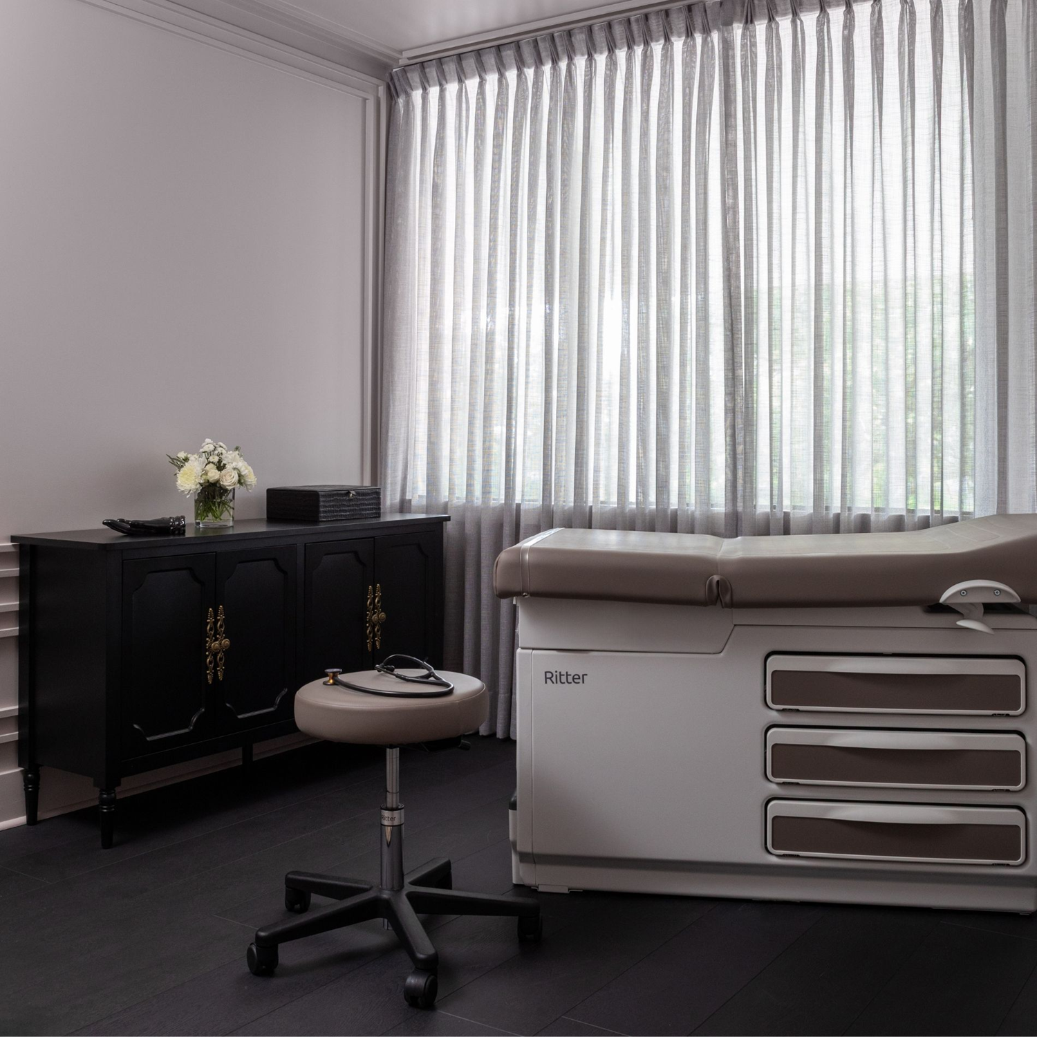Wellness clinic Houston treatment room with an exam table, clinician stool, black cabinet and soft sheer curtains.