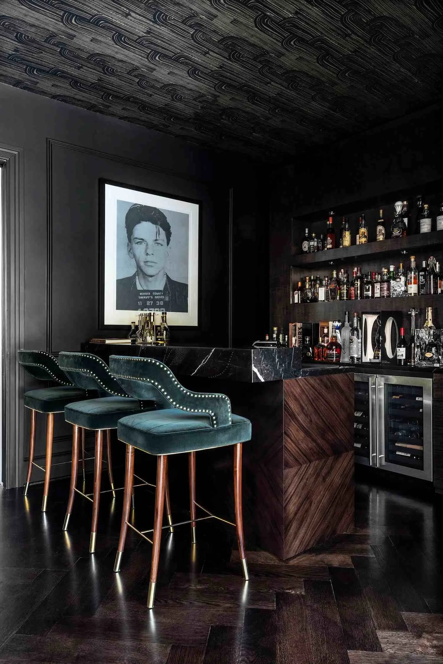 Dark home bar at the Saddlebranch II residence with green velvet stools, marble counter and portrait photography.