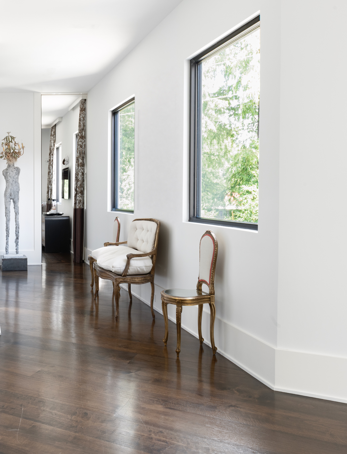 Modern luxury home in Houston hallway with dark wood floors, large windows, antique chairs and sculptural art.