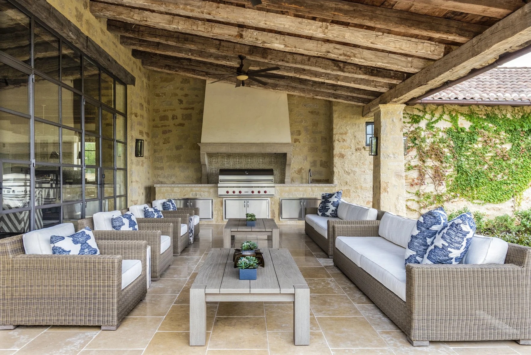 Modern ranch outdoor living area with reclaimed wood beams, stone walls, woven seating and an outdoor kitchen.