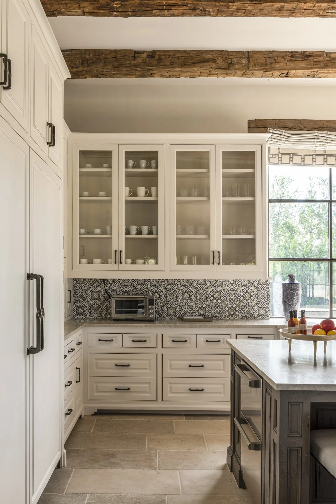 Modern ranch kitchen detail with glass-front white cabinets, patterned tile backsplash, stone flooring and reclaimed beams.