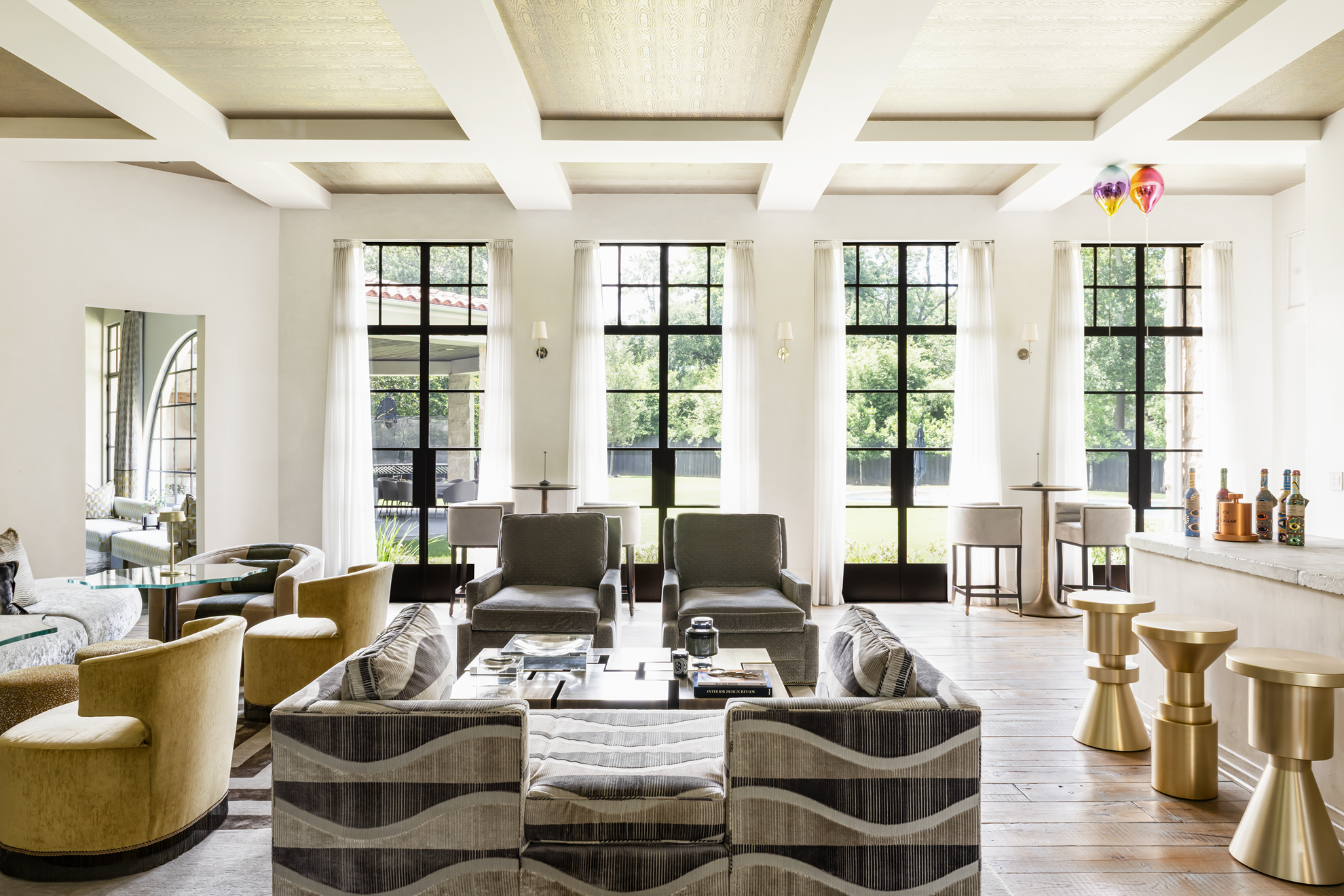 French country home in Houston open-plan lounge and bar with layered seating, brass stools and tall black-framed windows.