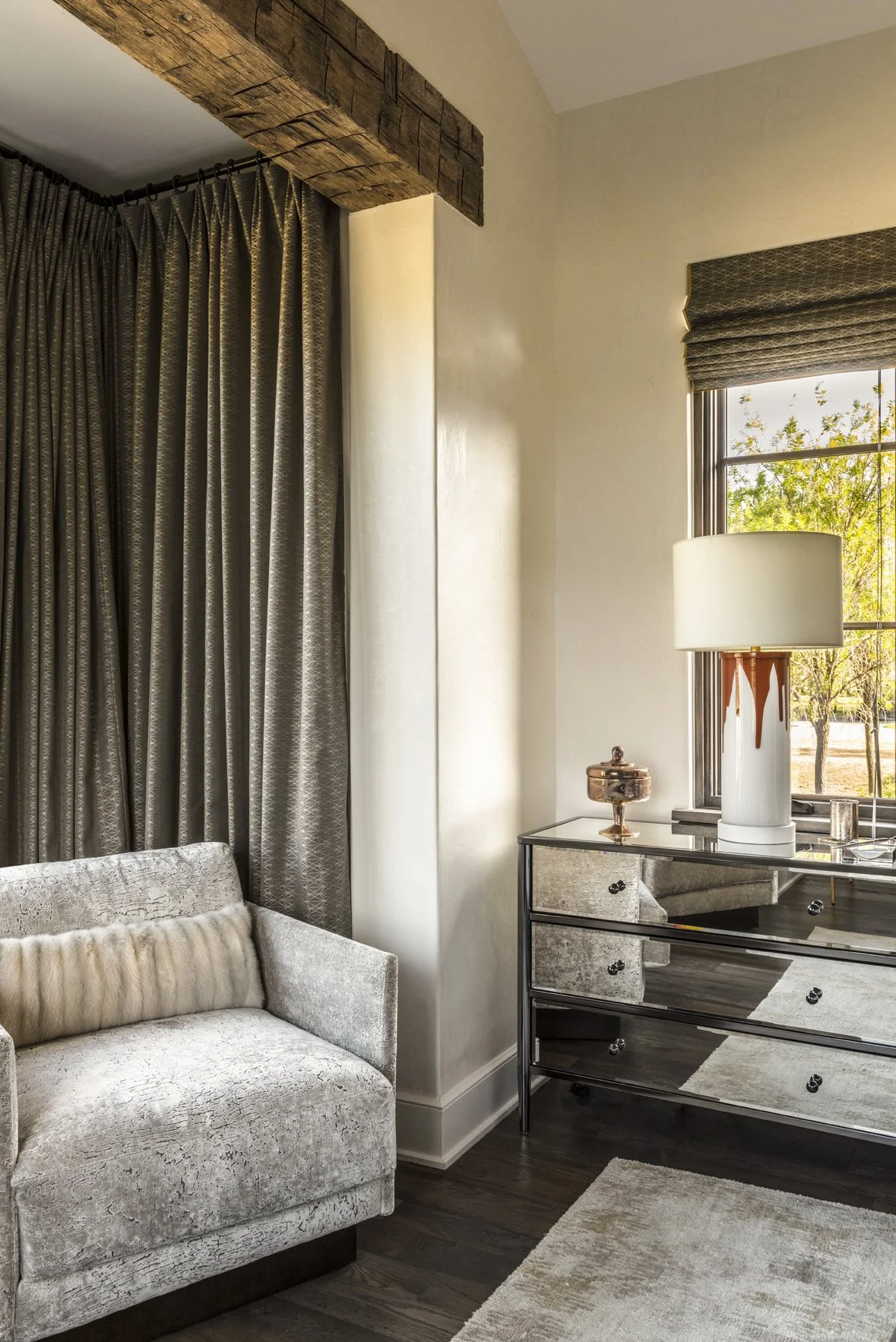 Modern ranch bedroom sitting area with a pale armchair, mirrored dresser, patterned drapery and reclaimed timber beam.