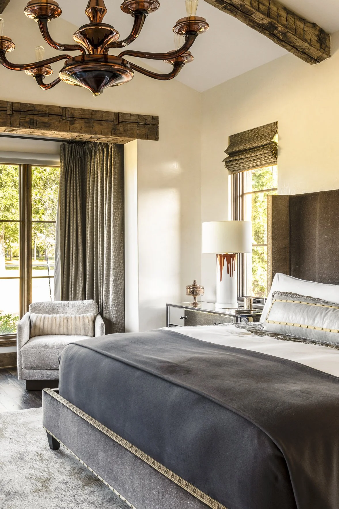 Luxury ranch bedroom interior with a gray upholstered bed, sculptural chandelier, mirrored nightstand and reclaimed wood beam.
