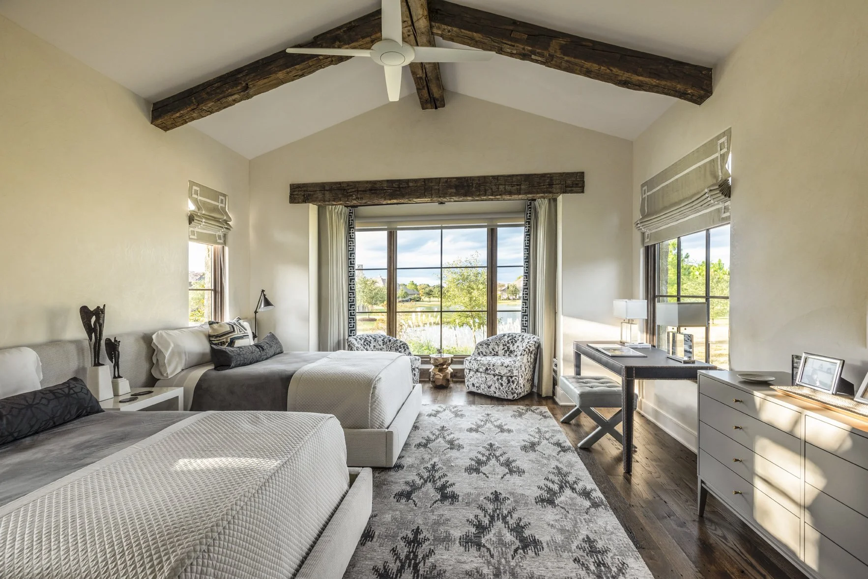 Modern ranch twin bedroom with two upholstered beds, reclaimed beams, patterned chairs, writing desk and a wide lake view.