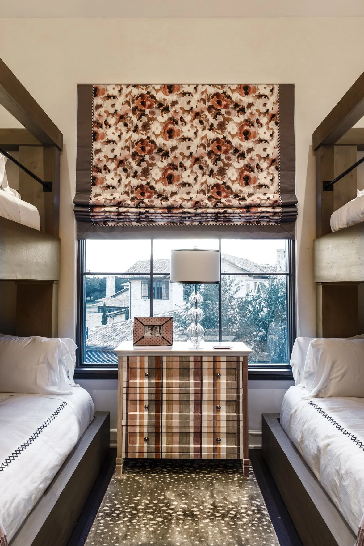 Modern ranch bunk room with built-in twin beds, a plaid chest, floral Roman shade and a large window between the beds.