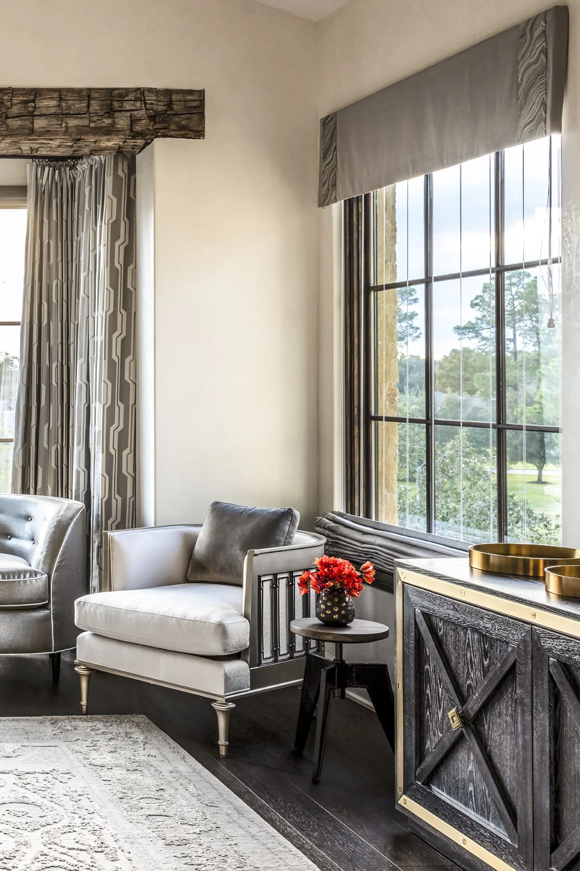 Luxury ranch bedroom sitting area with a tailored armchair, dark wood cabinet, red floral accent and large garden-facing windows.