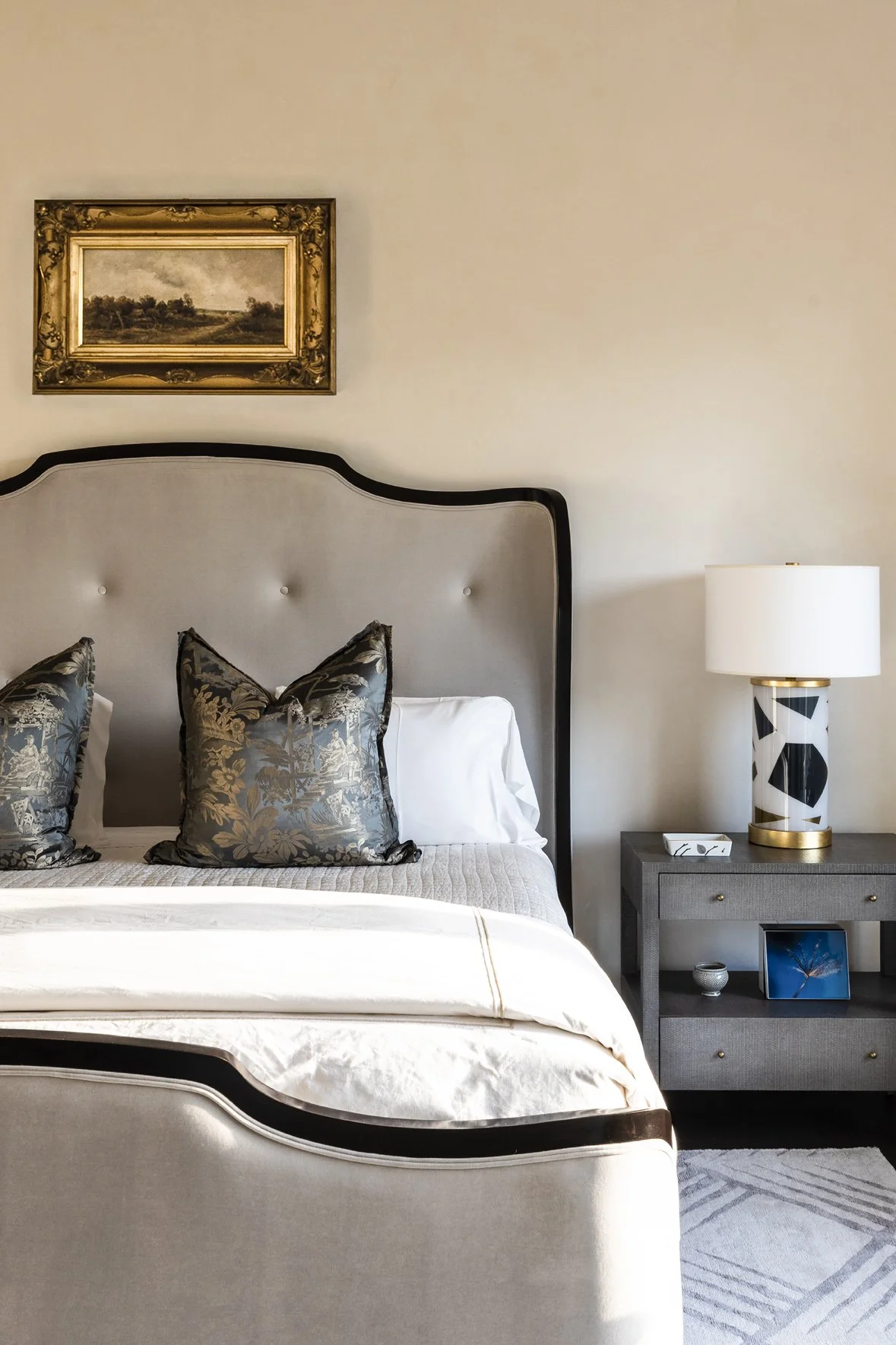 Luxury ranch bedroom detail with a curved upholstered headboard, patterned pillows, gray nightstand and graphic table lamp.
