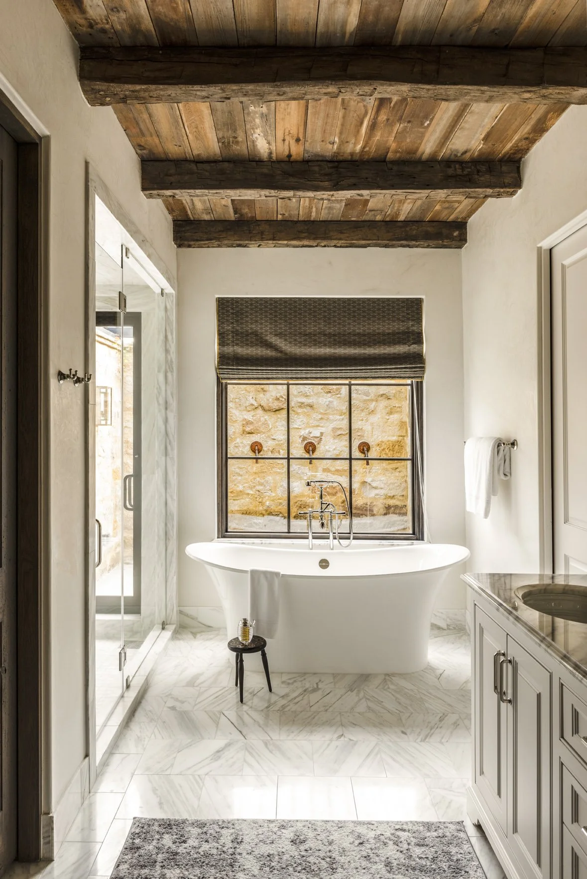 Luxury ranch bathroom interior with a freestanding tub, marble floor and vanity, black-framed mirror and reclaimed wood ceiling.