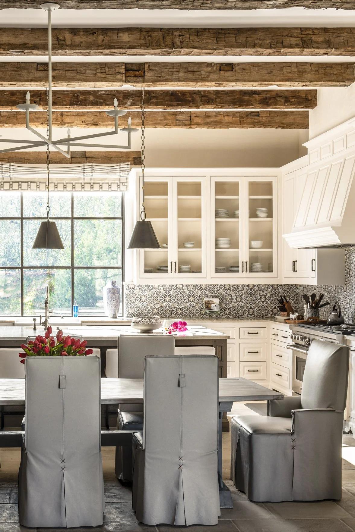 Modern ranch kitchen and dining area with white cabinetry, reclaimed wood beams, patterned tile and gray upholstered chairs.
