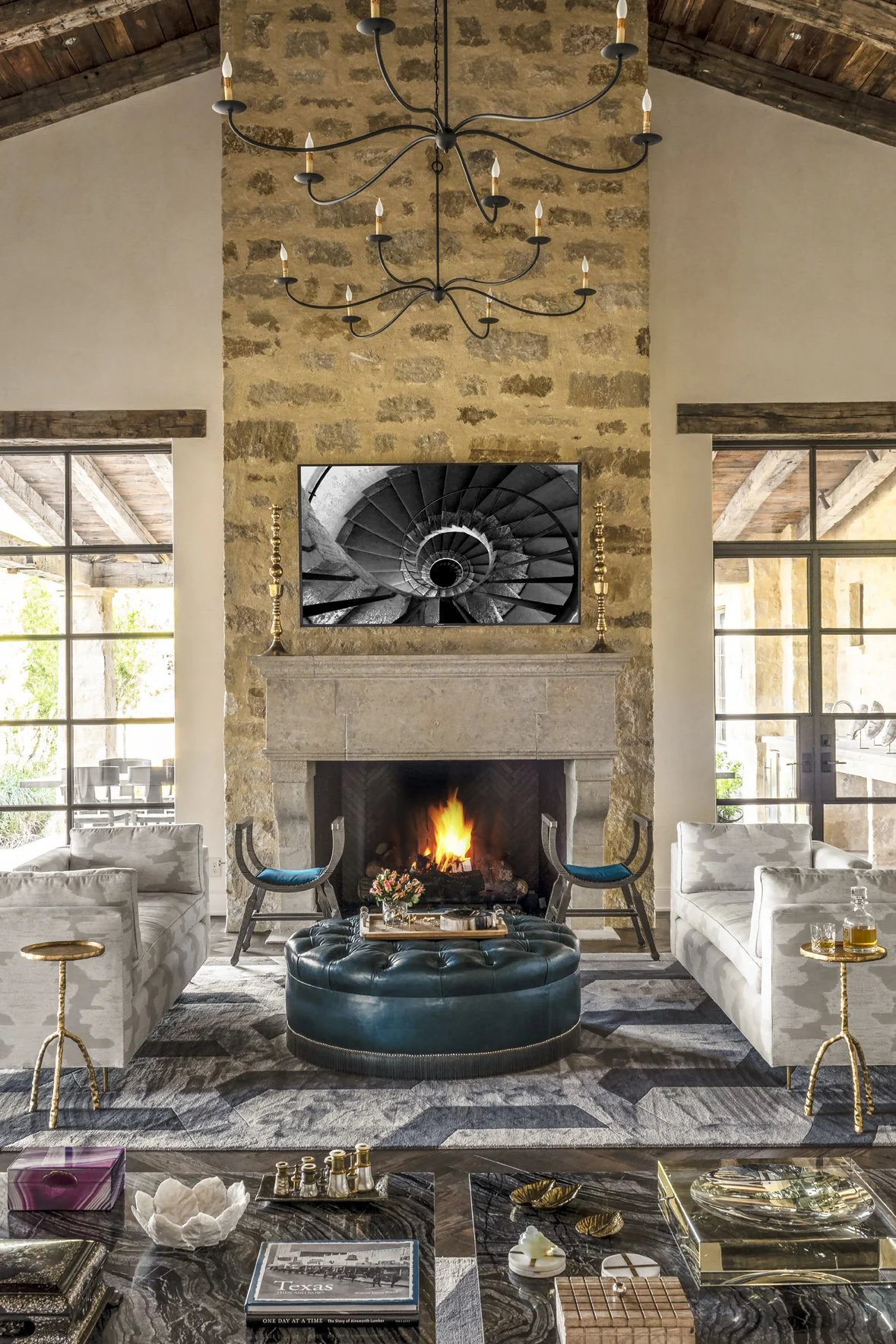 Texas ranch interior design in the great room with a tall stone fireplace, twin chandeliers and a blue tufted ottoman.