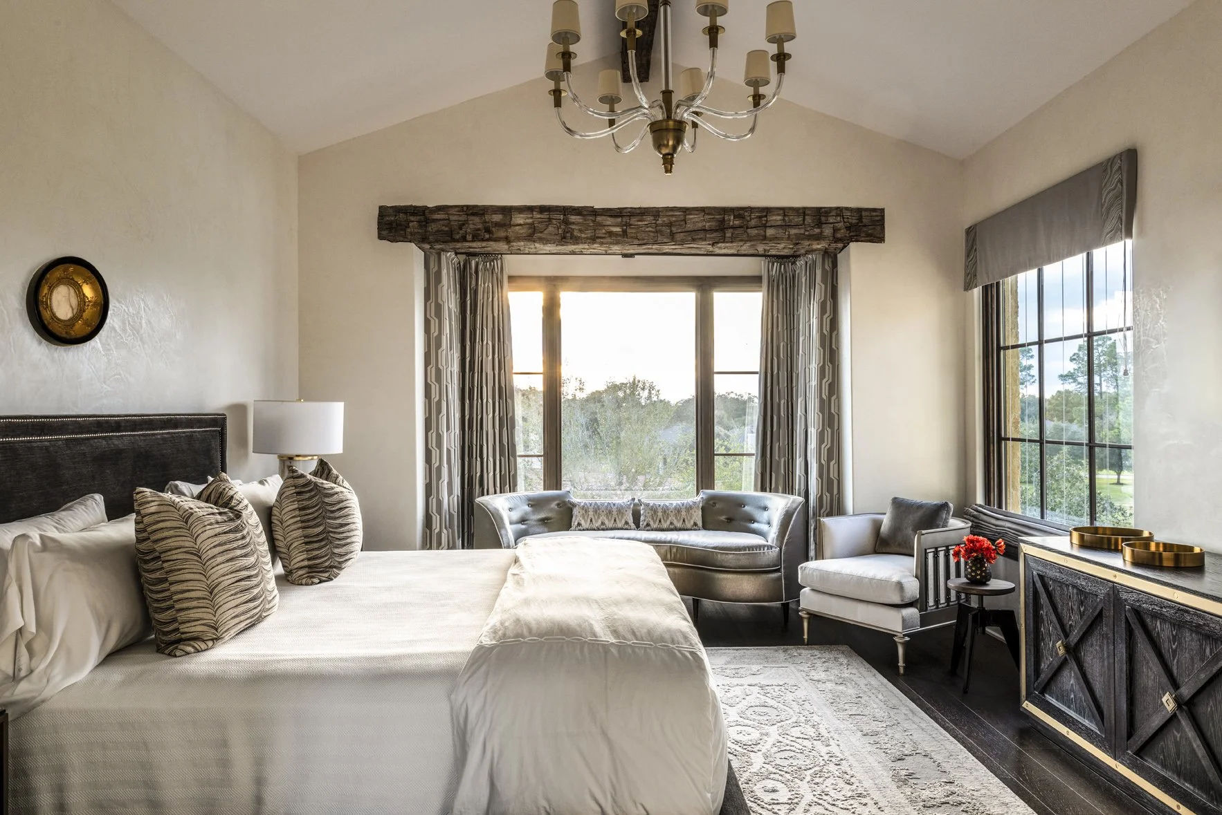 Modern ranch bedroom interior with a tailored bed, curved settee, armchair, reclaimed timber beam and dark wood cabinet.