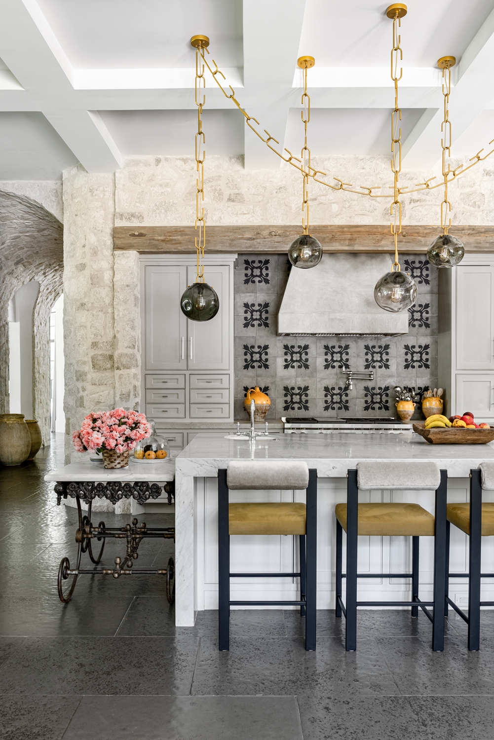 French country home in Houston kitchen with a marble island, mustard stools, patterned backsplash and stone walls.