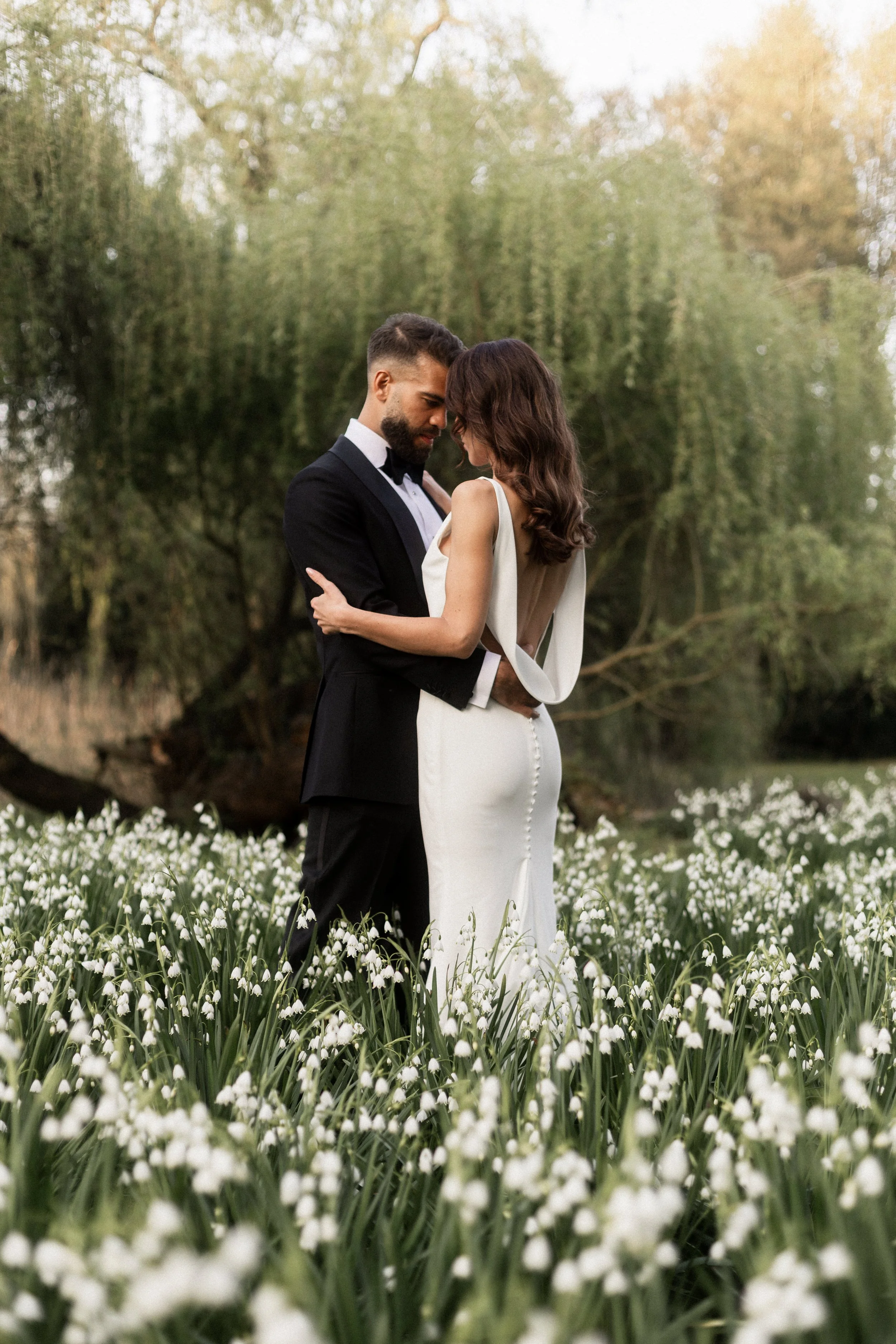 Un couple en tenue de mariage se tient dans un champ de jonquilles, avec des arbres en arrière-plan, lors d'une séance photo en plein air.