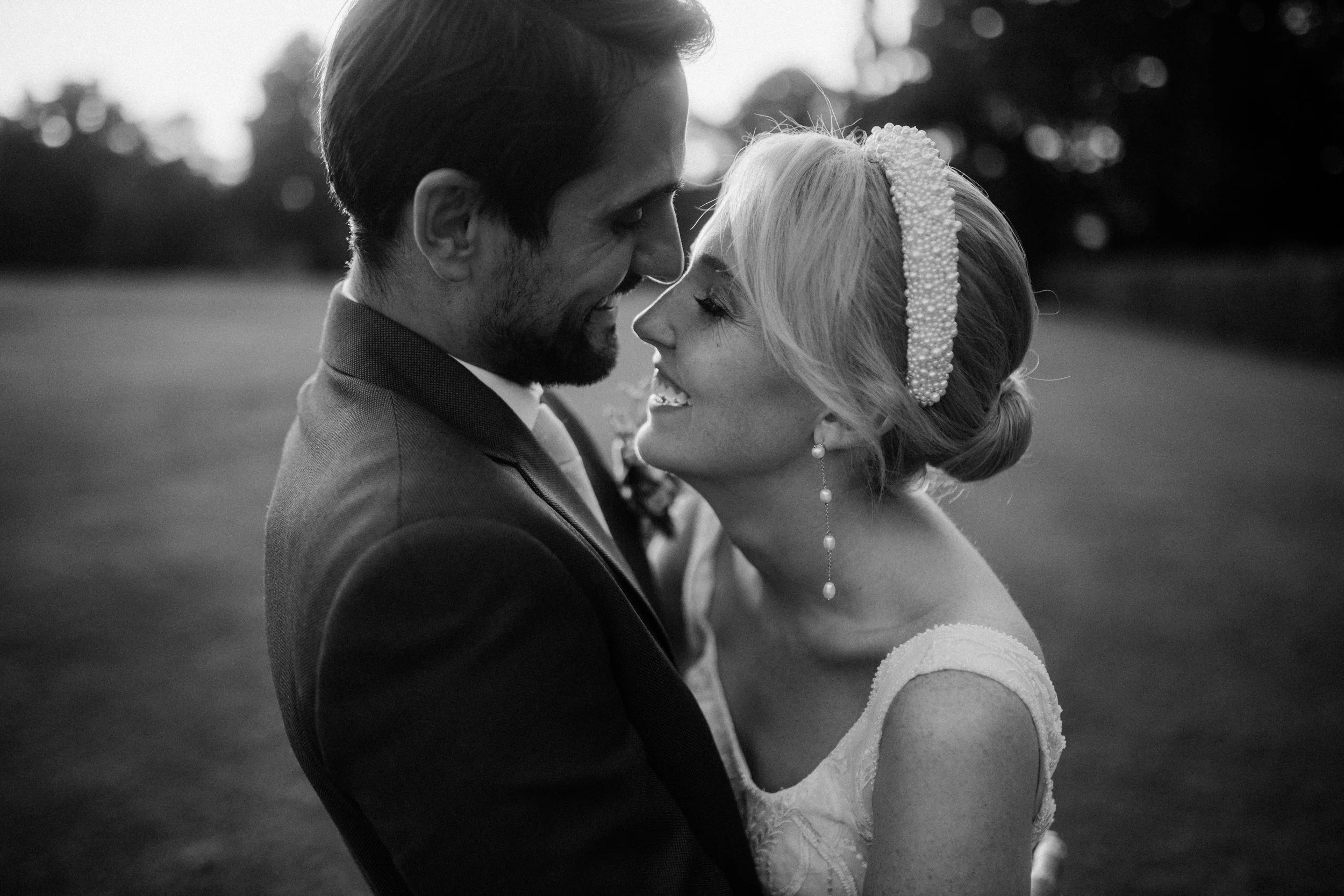 Un couple marié, un homme et une femme, se regardent tendrement en noir et blanc lors de leur mariage. La femme porte une coiffure élégante avec un bandeau de perles, une robe de mariage et des boucles d'oreilles pendantes. L'homme porte un costume sombre et une cravate. Ils semblent très heureux, dans un cadre naturel avec des arbres en arrière-plan.