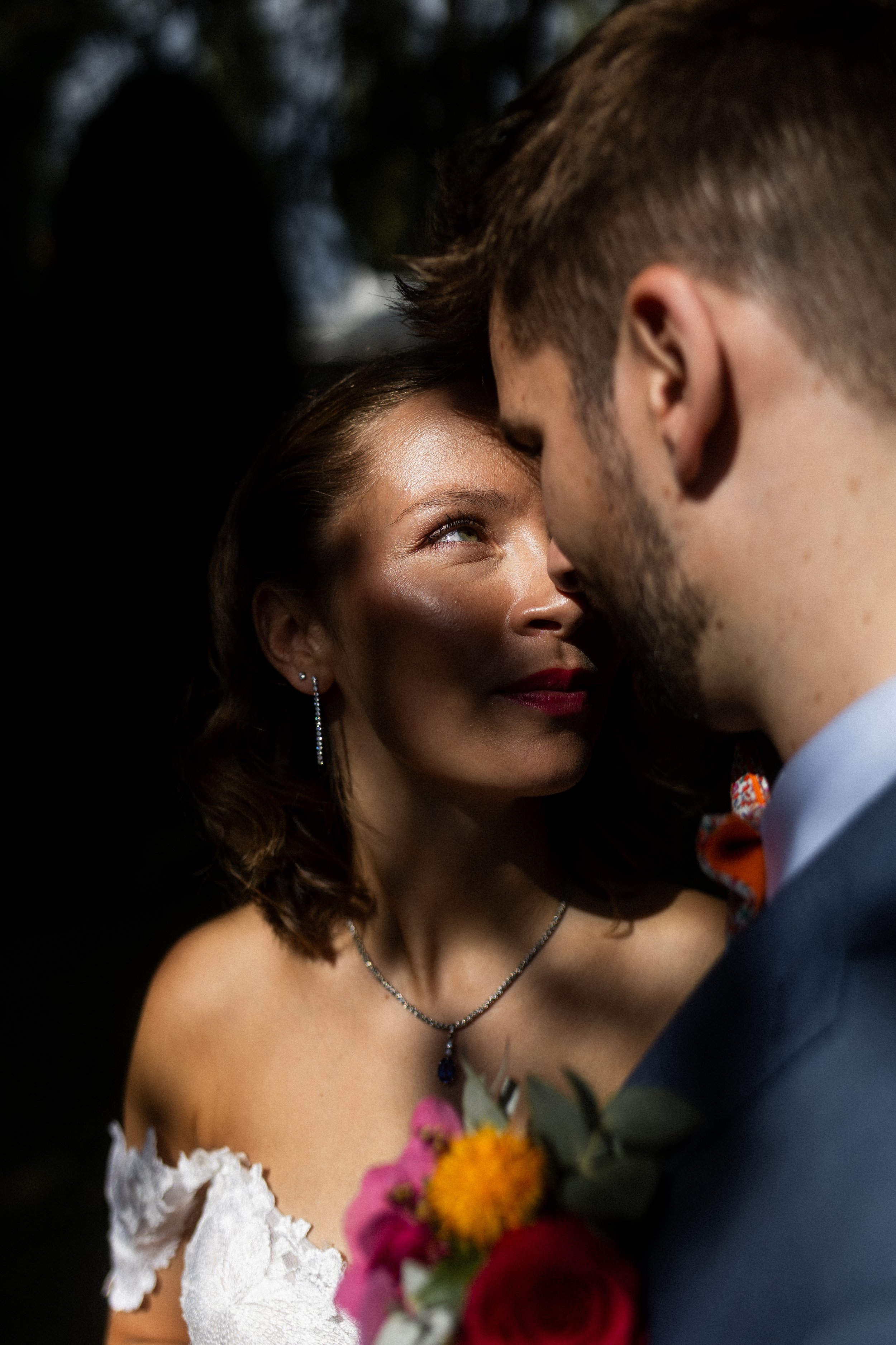Un couple en tenue élégante, probablement lors d'un mariage, se regarde avec tendresse. La femme porte une robe blanche avec des détails en dentelle, des bijoux, et a les cheveux bouclés. La scène est romantique et intime.