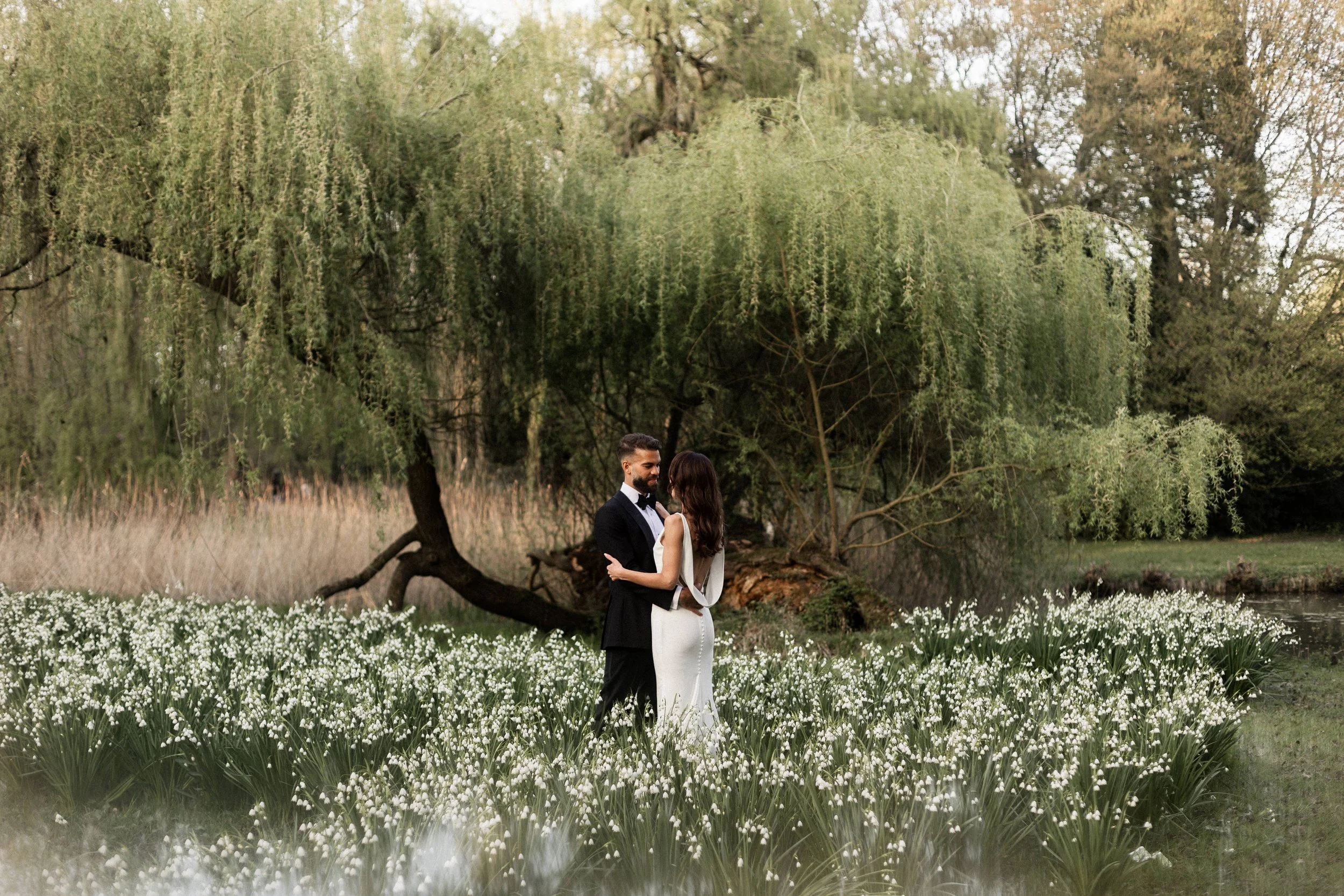 Un couple marié, un homme en smoking et une femme en robe blanche, se tiennent la main dans un cadre naturel avec des arbres et une étendue de fleurs blanches au sol.