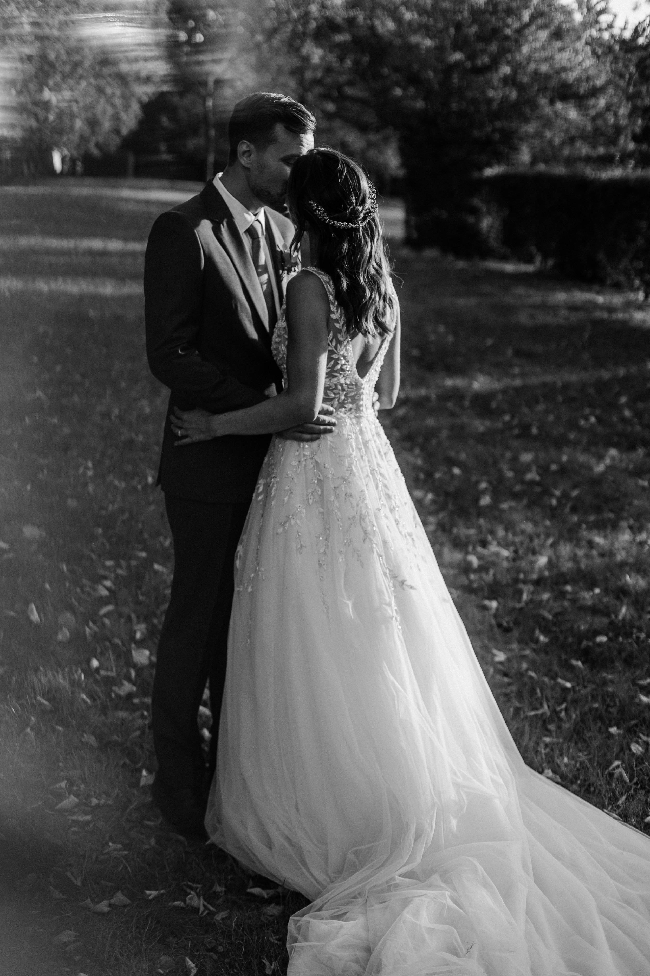 Un couple en habits de mariage s'embrasse dans un parc, en noir et blanc.