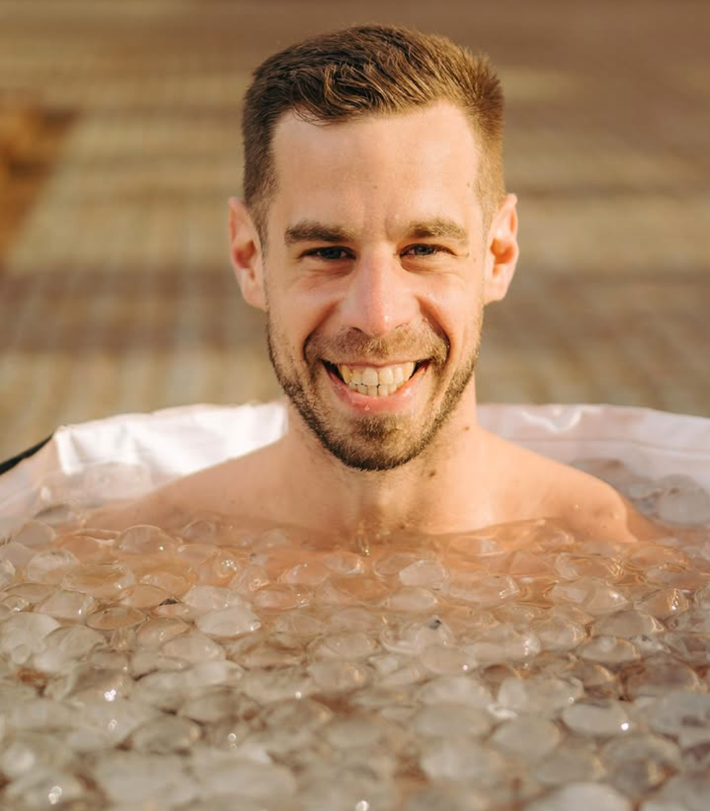 Man die lachend in een bad met ijsklonten ligt, met een achtergrond van een houten vloer.