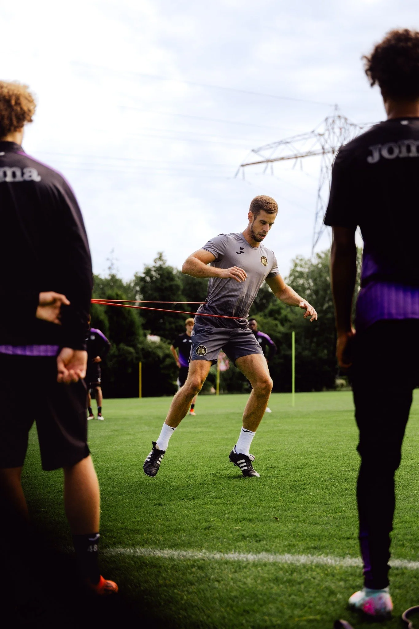 Een man draagt een trainingspak en doet een trainingsessie met weerstandsbanden op een grasveld met andere mensen op de achtergrond.