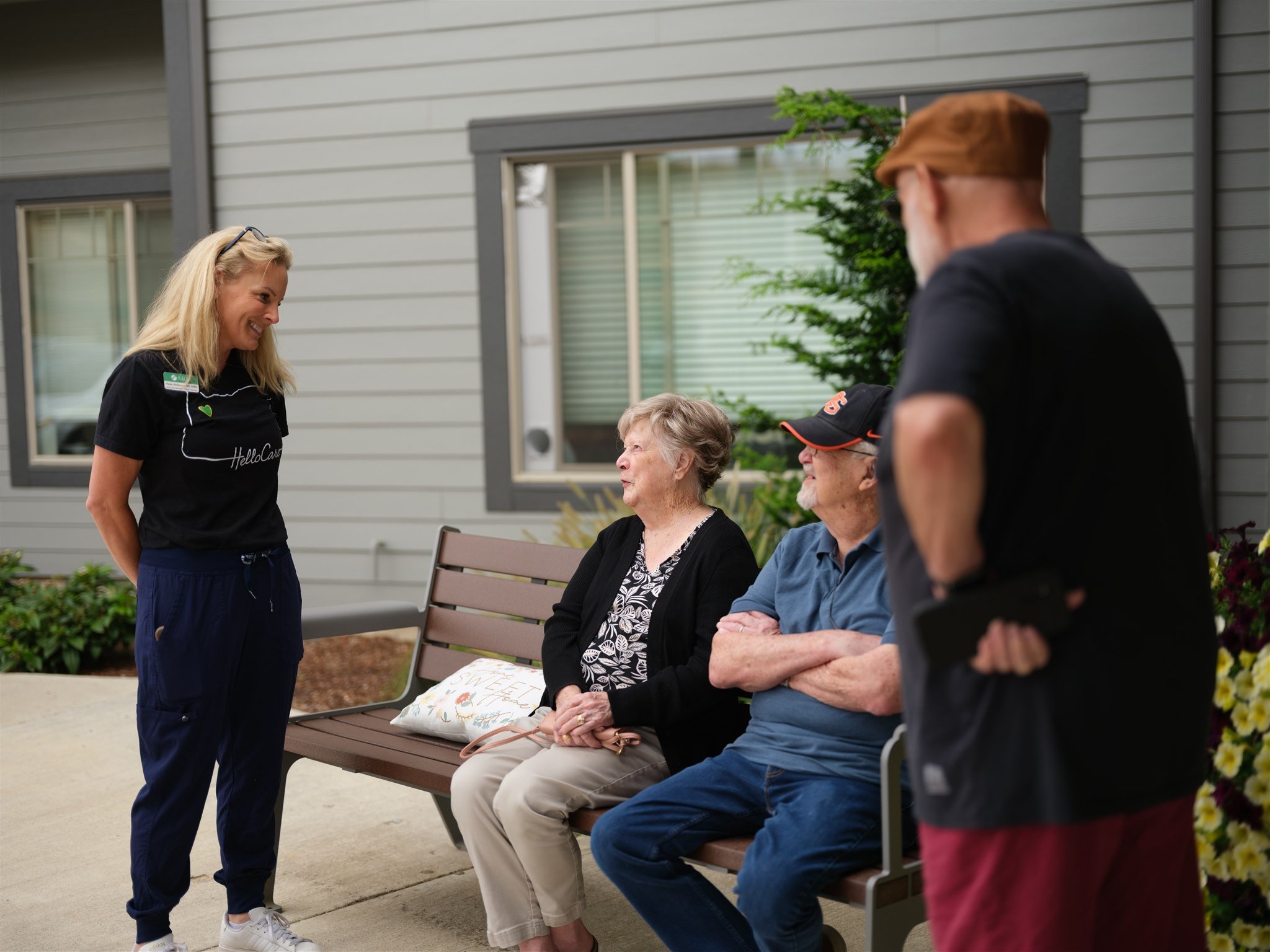 A young woman is standing and smiling at three older adults sitting on a bench outside a house, engaged in a conversation.