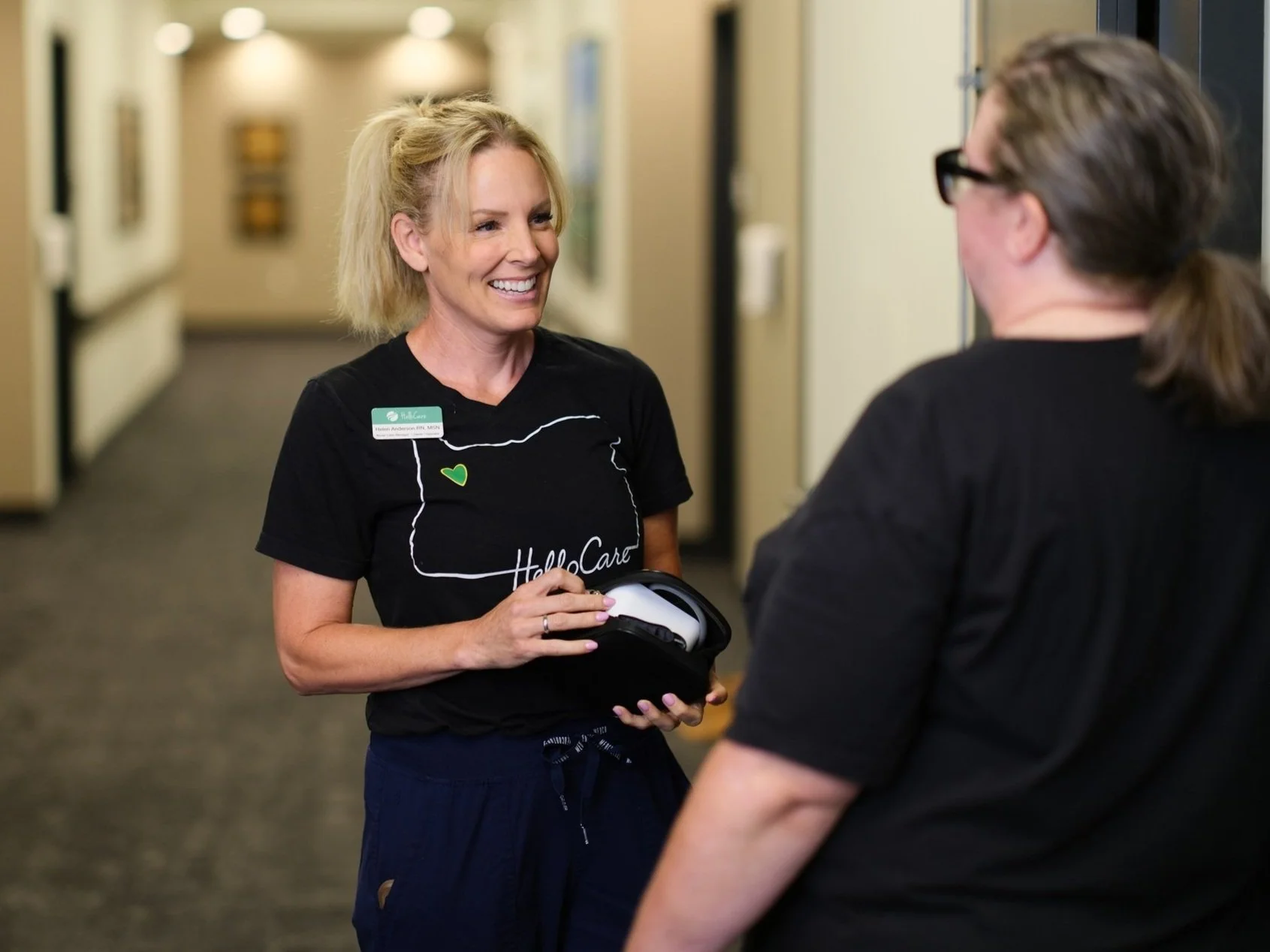 Two women having a conversation in a hallway, one woman is smiling while holding a mask, and the other woman is seen from the back wearing glasses.