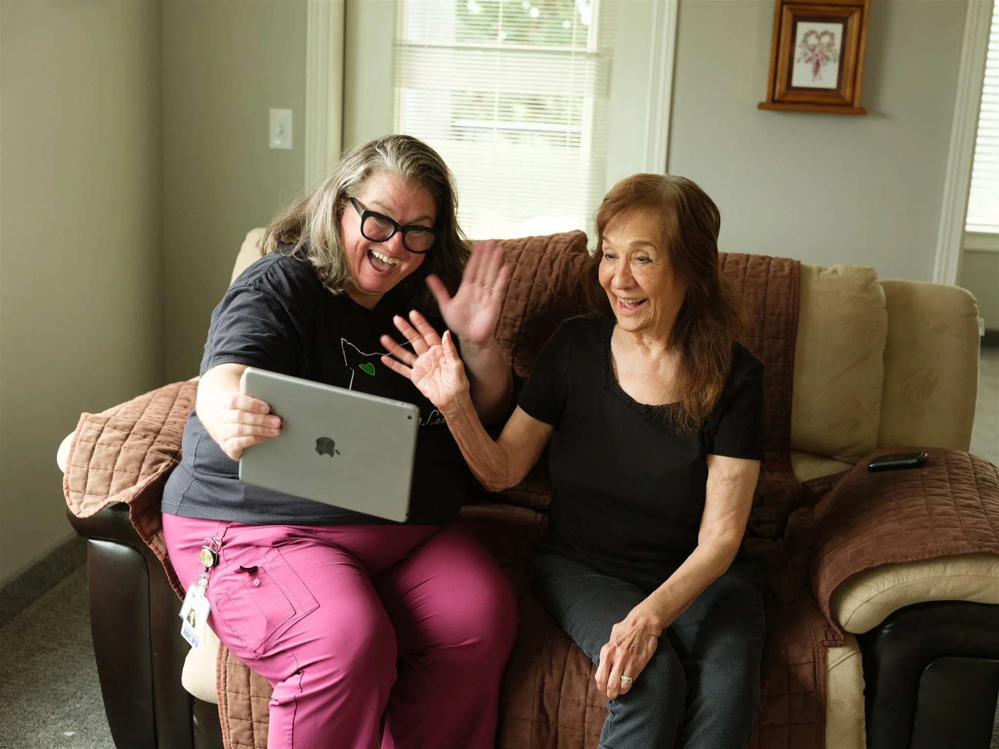 Two women sitting on a couch looking at a tablet and laughing. One woman wears glasses and pink pants, the other woman wears a black shirt and gray pants. They appear to be in a living room.