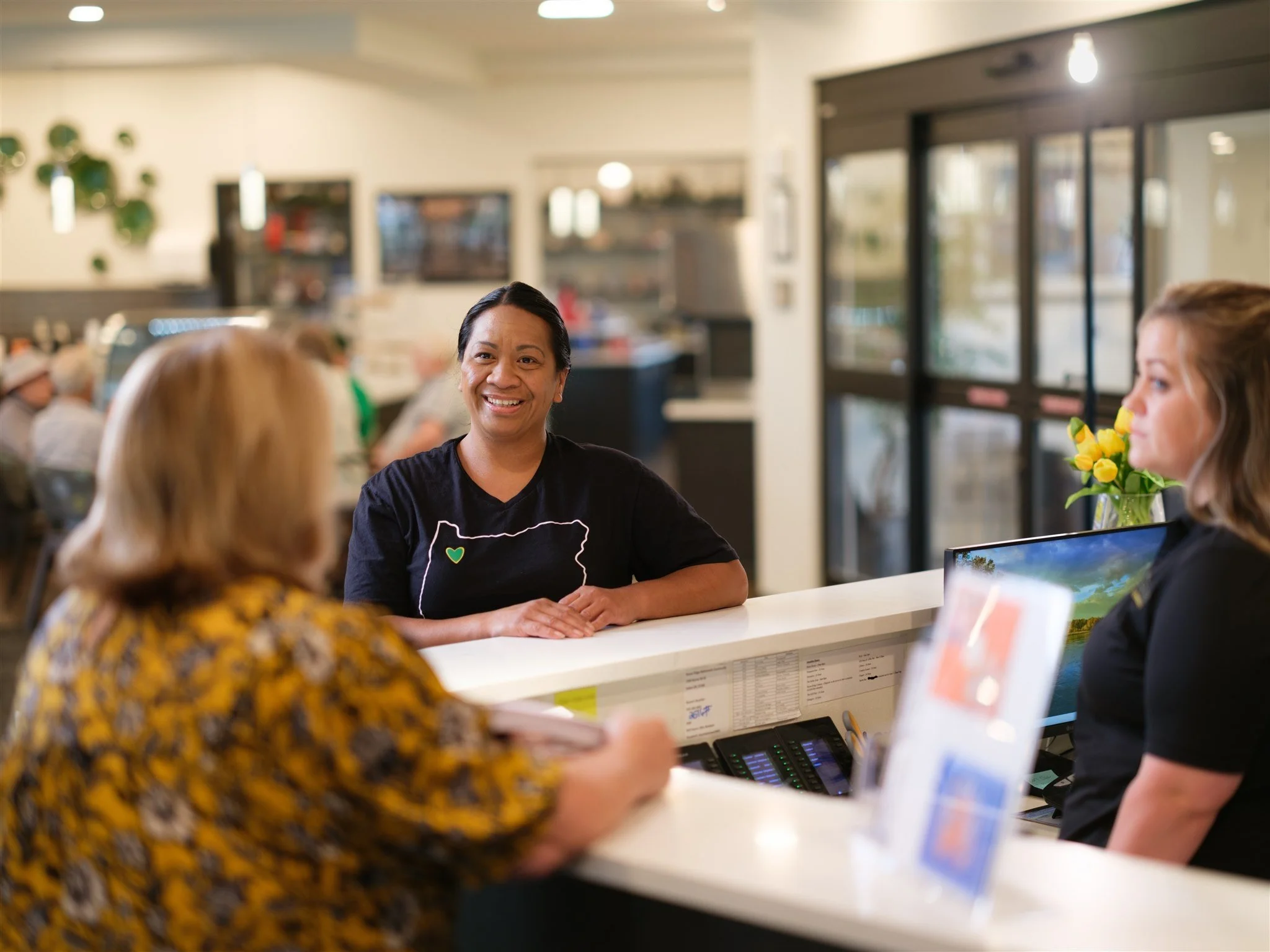 A smiling woman at the hotel reception desk speaking with two women, one with blonde hair in a yellow patterned blouse, and another with brown hair in a black shirt with a computer monitor, flowers, and a menu display on the counter.