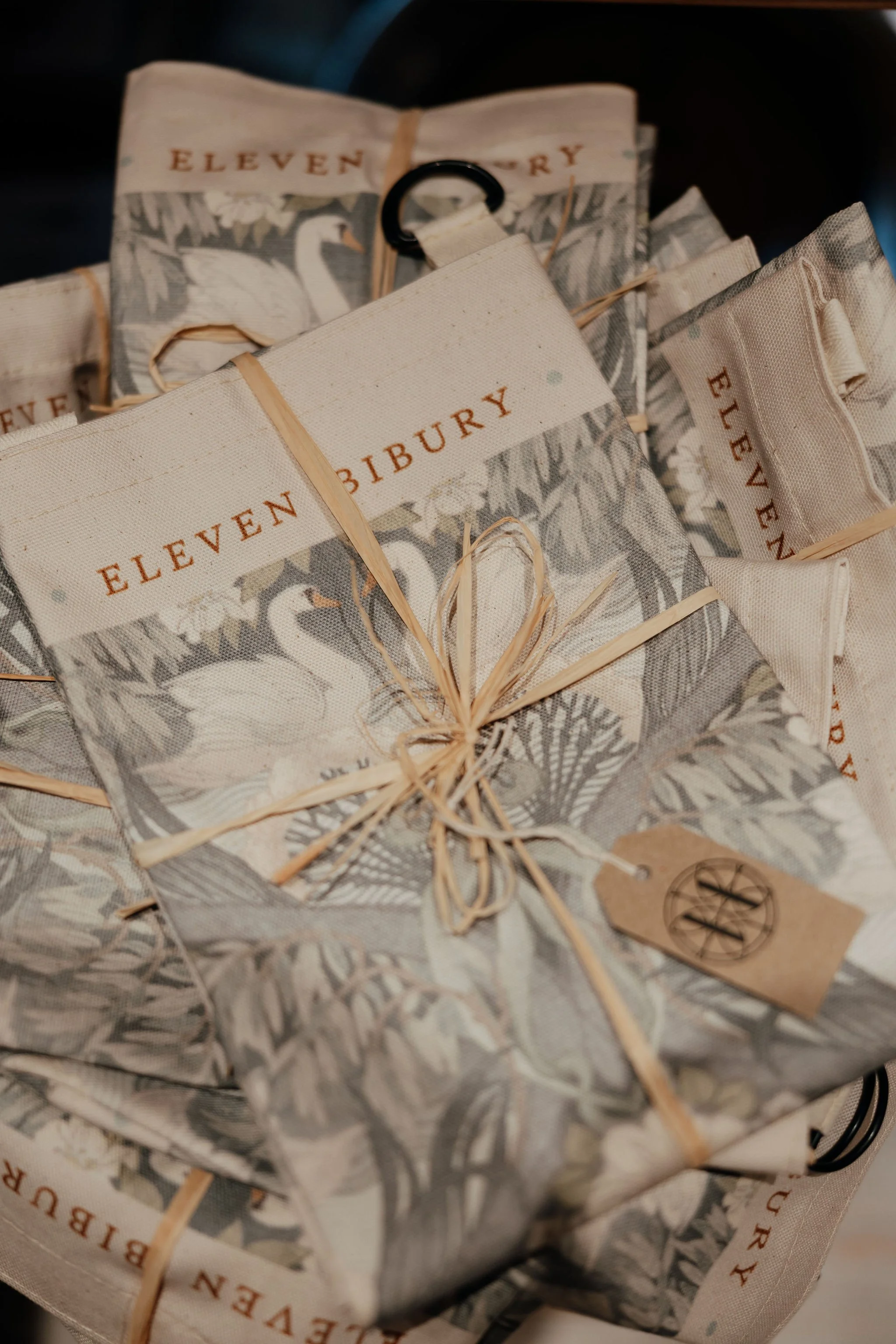 Several packages wrapped in botanical-patterned paper with the words "Eleven Bury" printed on them, tied with light brown raffia string, and a small tag with an emblem.