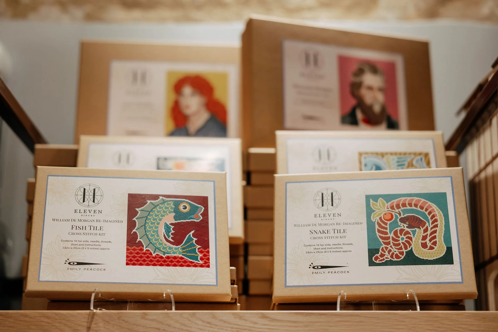 Two embroidery cross-stitch kits titled 'Fish Tile' and 'Snake Tile' displayed on a wooden shelf with illustrations of a fish and a snake, respectively, each in colorful square designs.