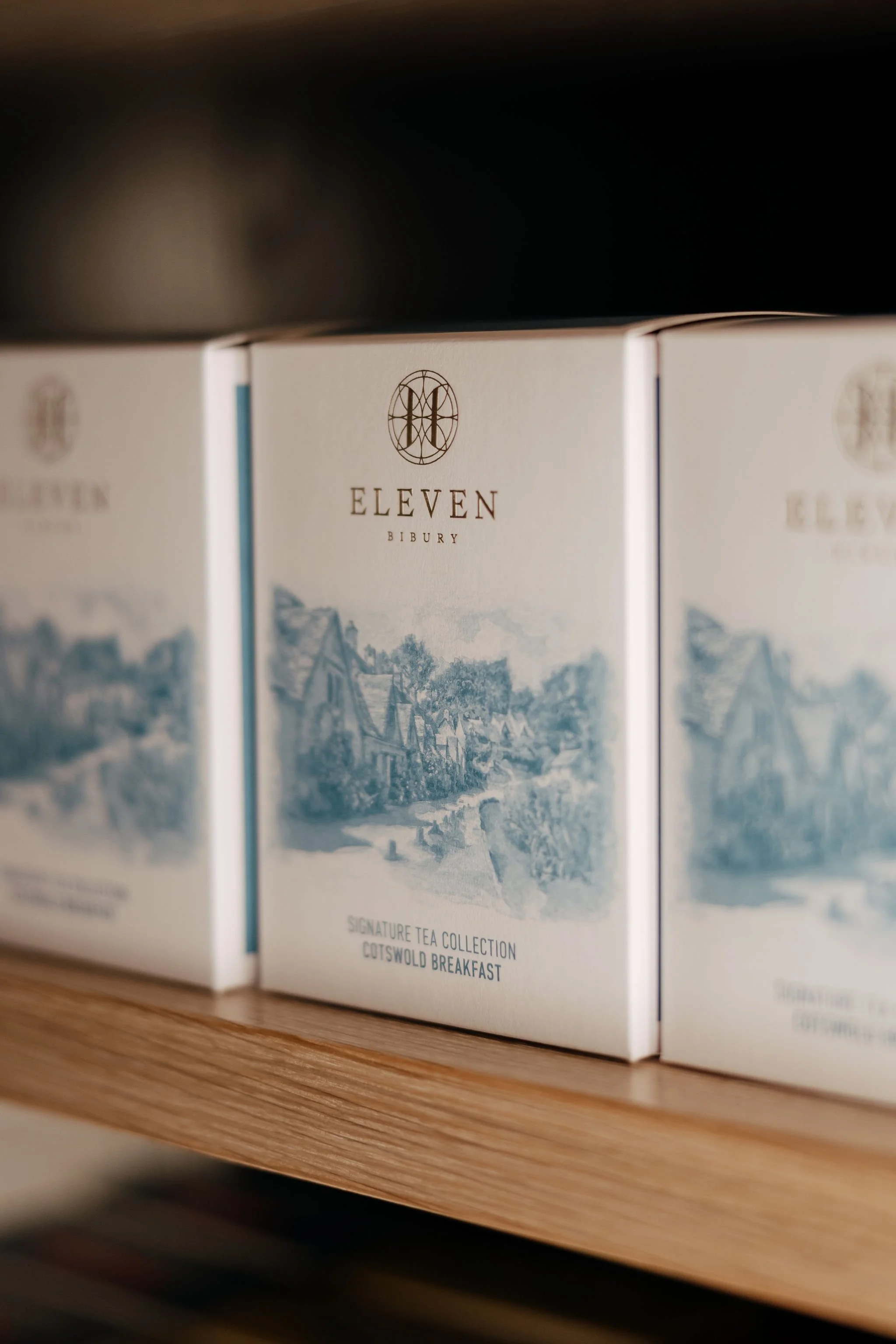Boxes of Eleven Bibury signature tea collection on a wooden shelf, with a scenic village illustration on the packaging.