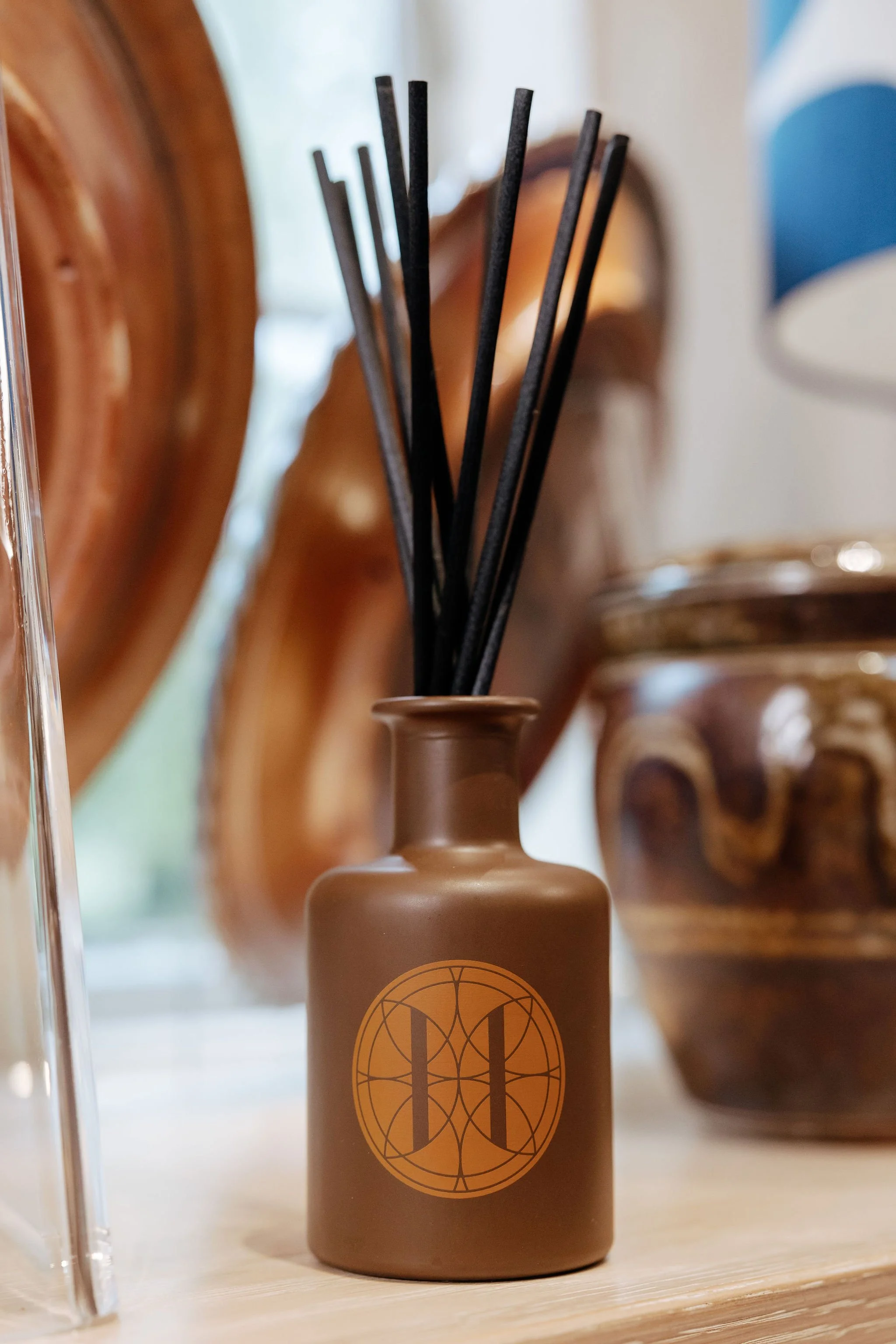 A small brown vase with a black geometric design, holding black reed diffusers, placed on a wooden surface with decorative wooden objects in the blurry background.