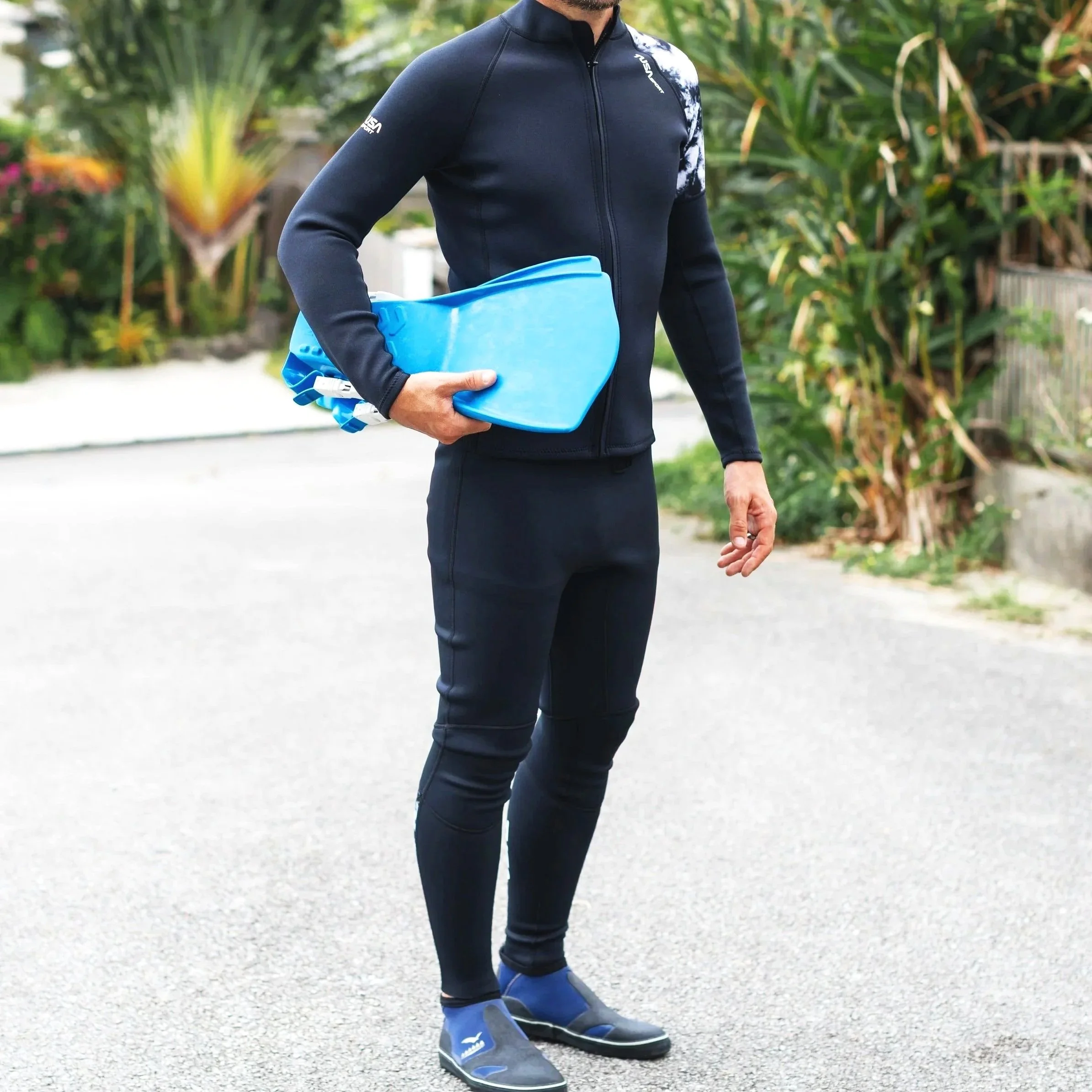 A person wearing a black athletic jacket with branding on the sleeves and chest, black pants, and black and blue athletic shoes, holding a blue item in their hand, standing outdoors on a paved surface with green plants and trees in the background.