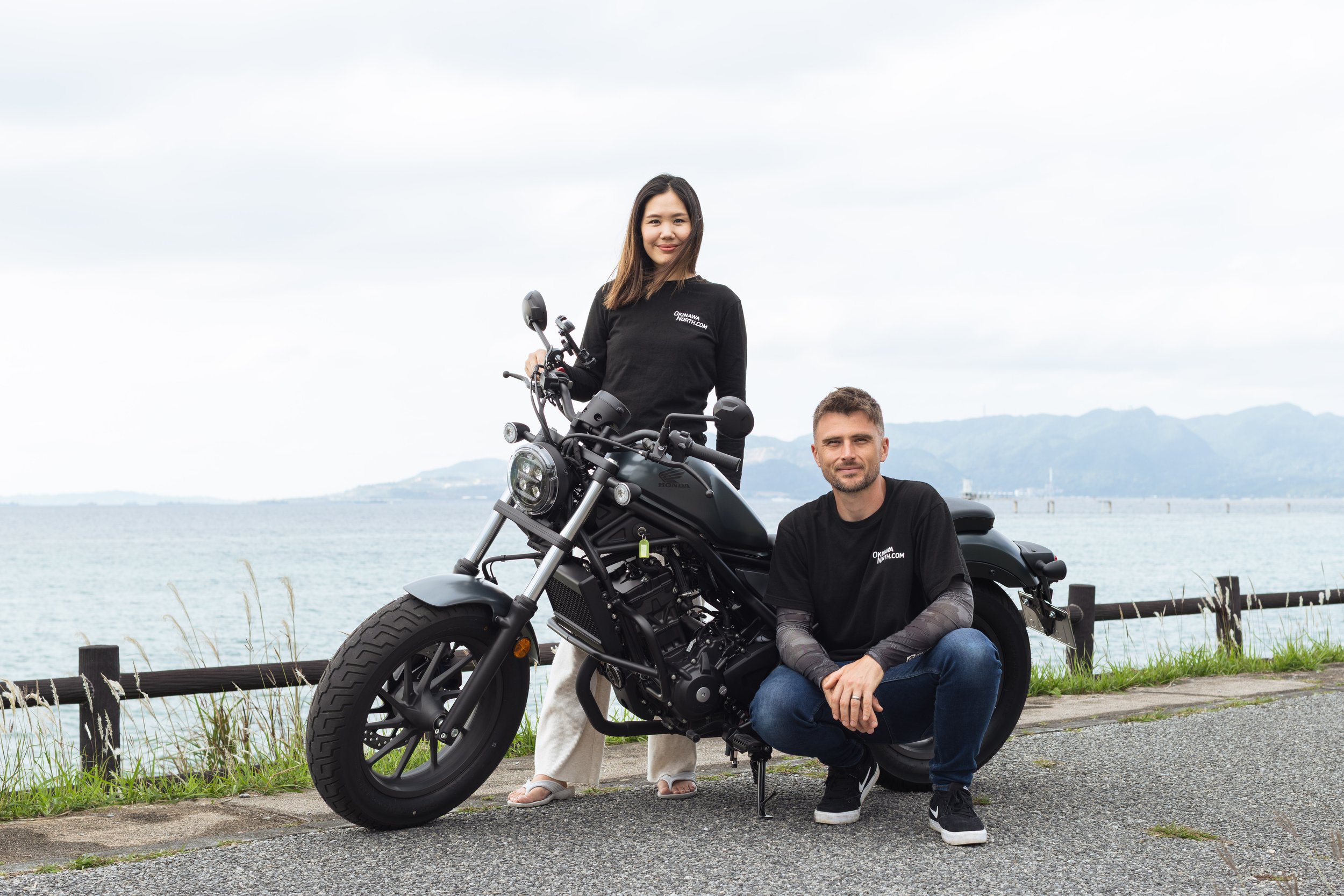 A woman standing next to a man sitting on a motorcycle near a body of water with mountains in the background. The woman is wearing a black shirt and white pants, while the man is wearing a black shirt and jeans. Both are smiling and looking at the camera.