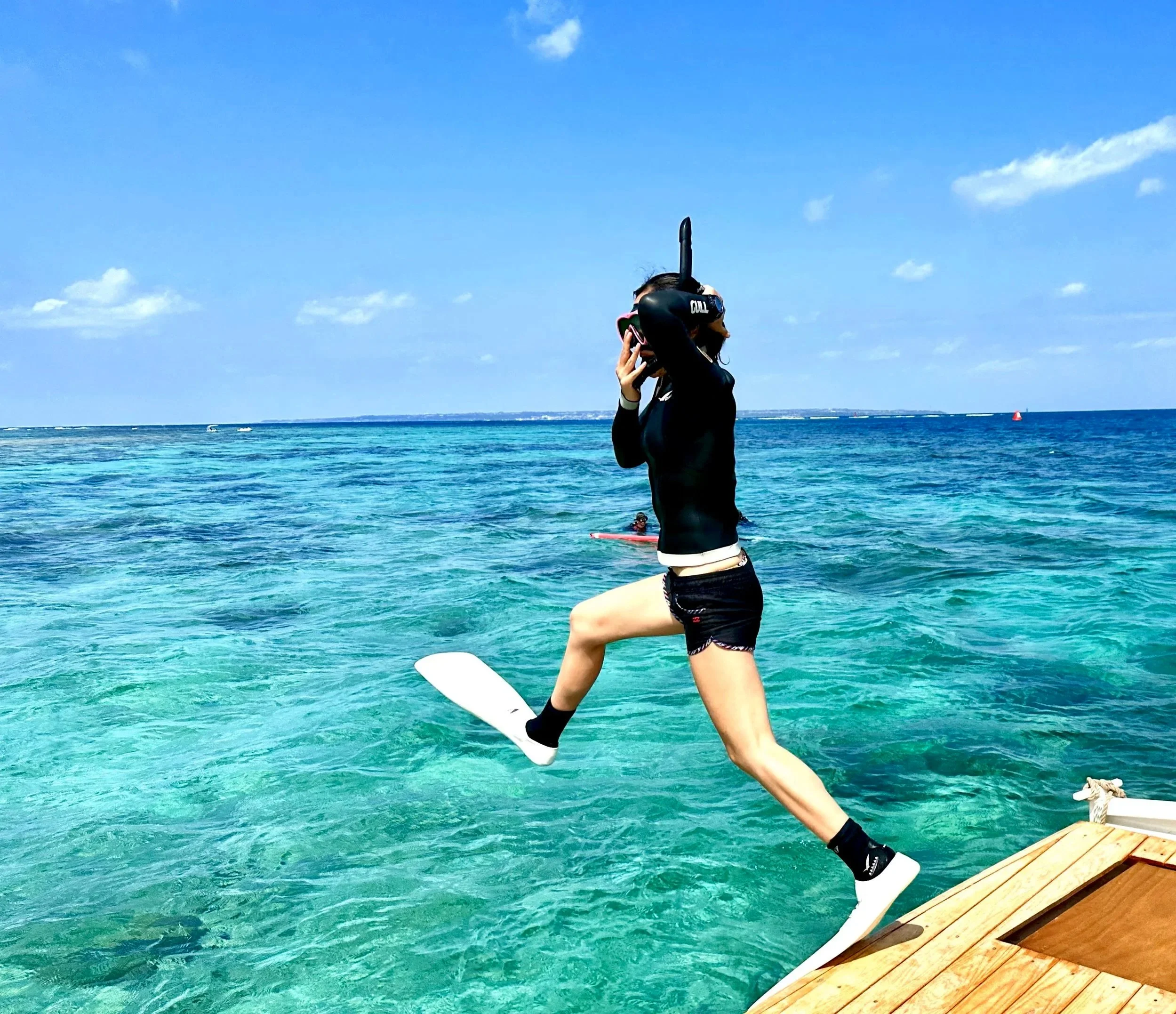 A person in a wetsuit jumping off a dock into clear blue water, holding a snorkeling mask, with the ocean and sky in the background.