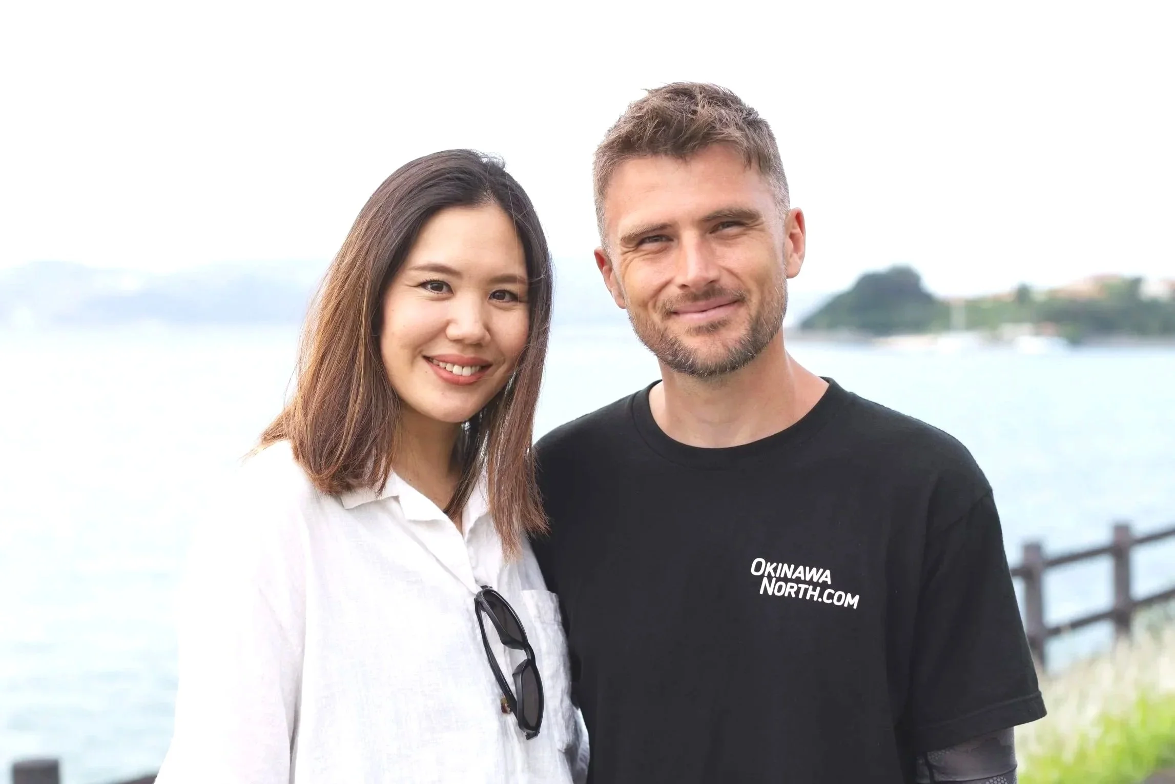 A man and a woman smiling outdoors with water and greenery in the background.