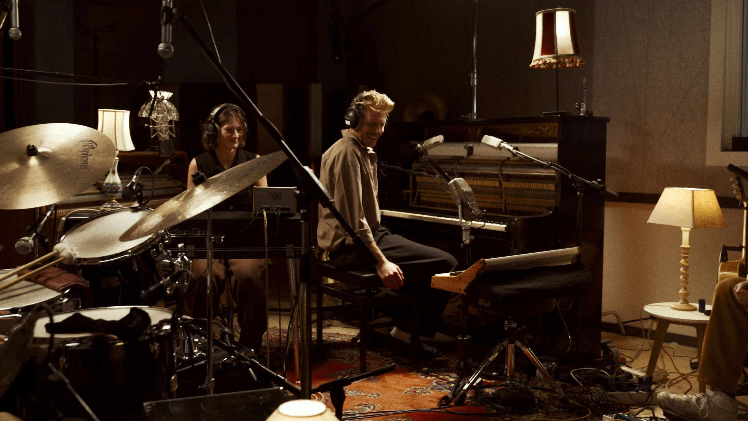 Musicians recording in a studio with drums, piano, microphones, and warm lighting.