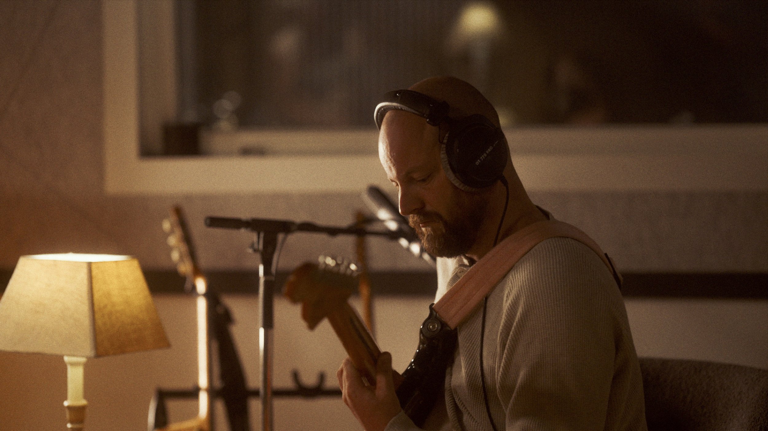 A man with a beard and bald head wearing headphones, playing an electric guitar in a dimly lit room with a lamp and musical equipment in the background.
