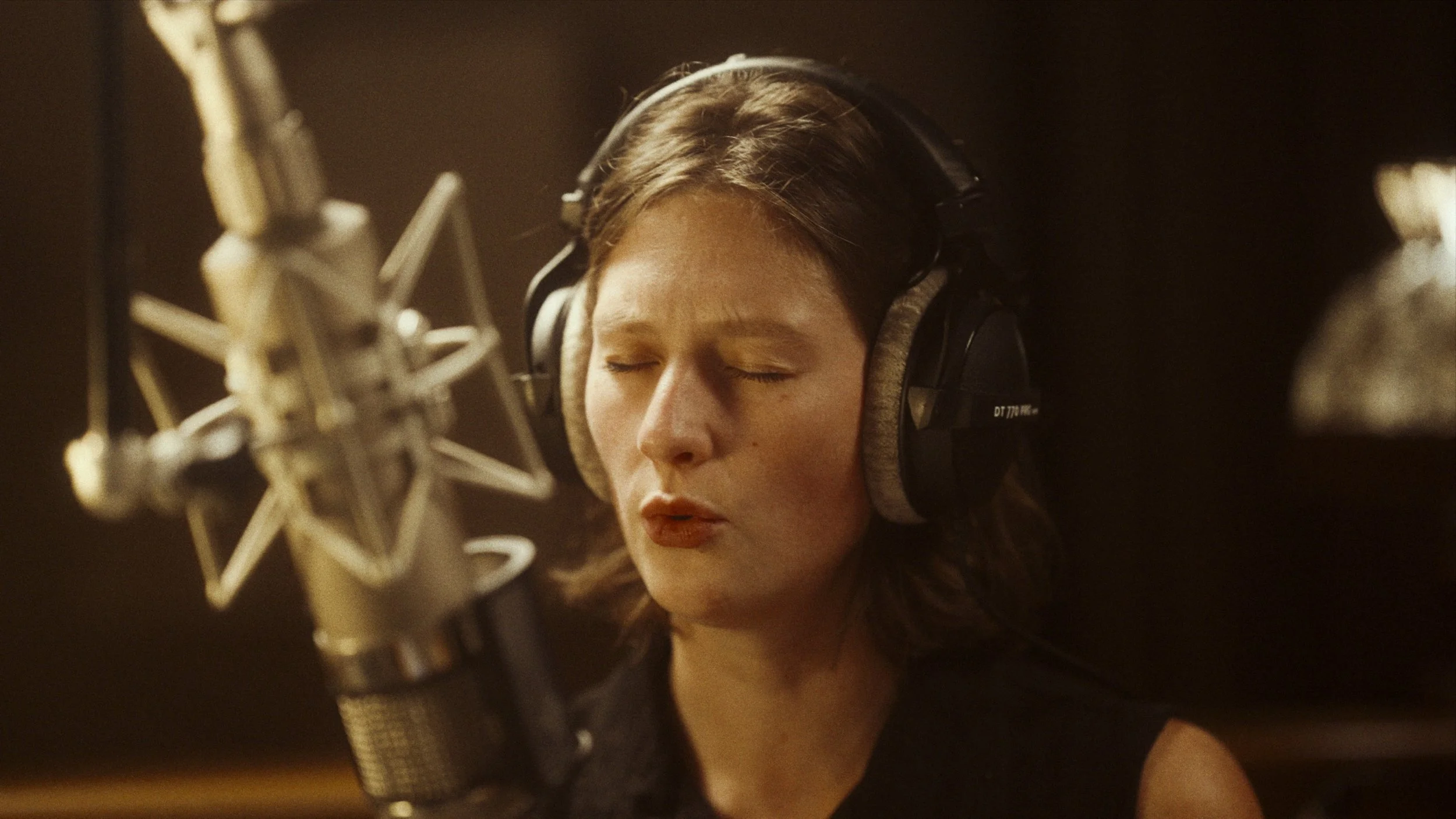 A woman with brown hair wearing headphones singing into a microphone in a recording studio.