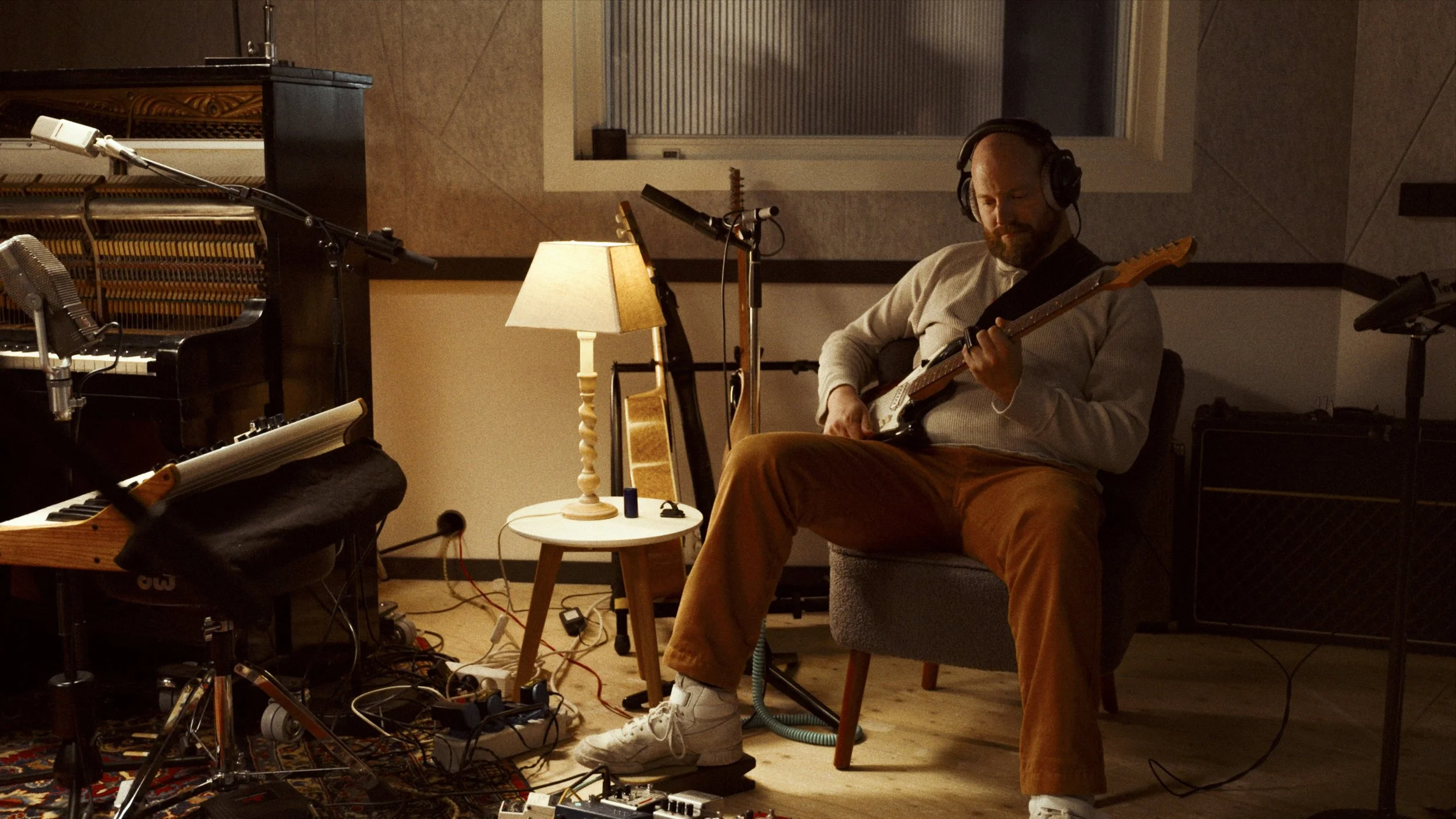 A man wearing a beige sweater and brown pants plays an electric guitar in a recording studio, surrounded by microphones, a grand piano, a lamp, and audio equipment.